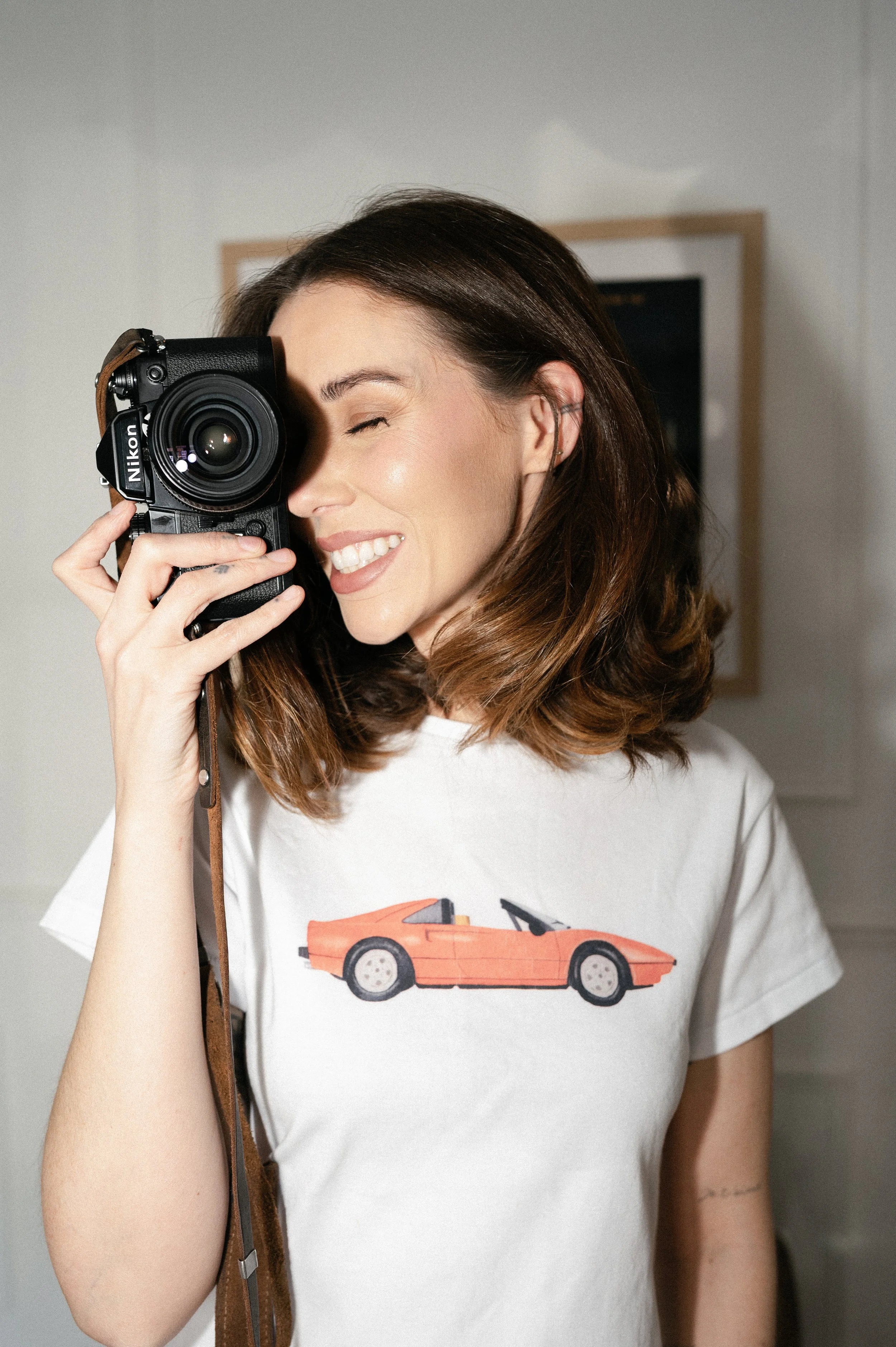 A woman with brown hair smiling and holding a Nikon camera to her face, with her eye closed, in an indoor setting.