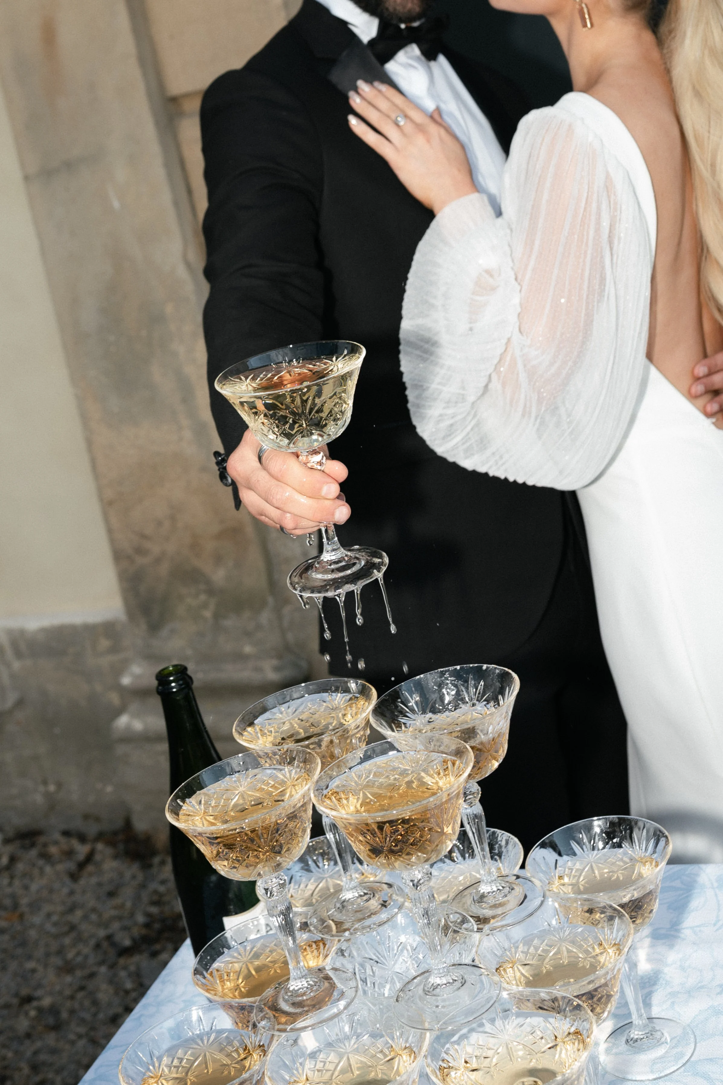A couple in formal wedding attire celebrating with champagne glasses, with champagne cascading from one glass into others on a table.