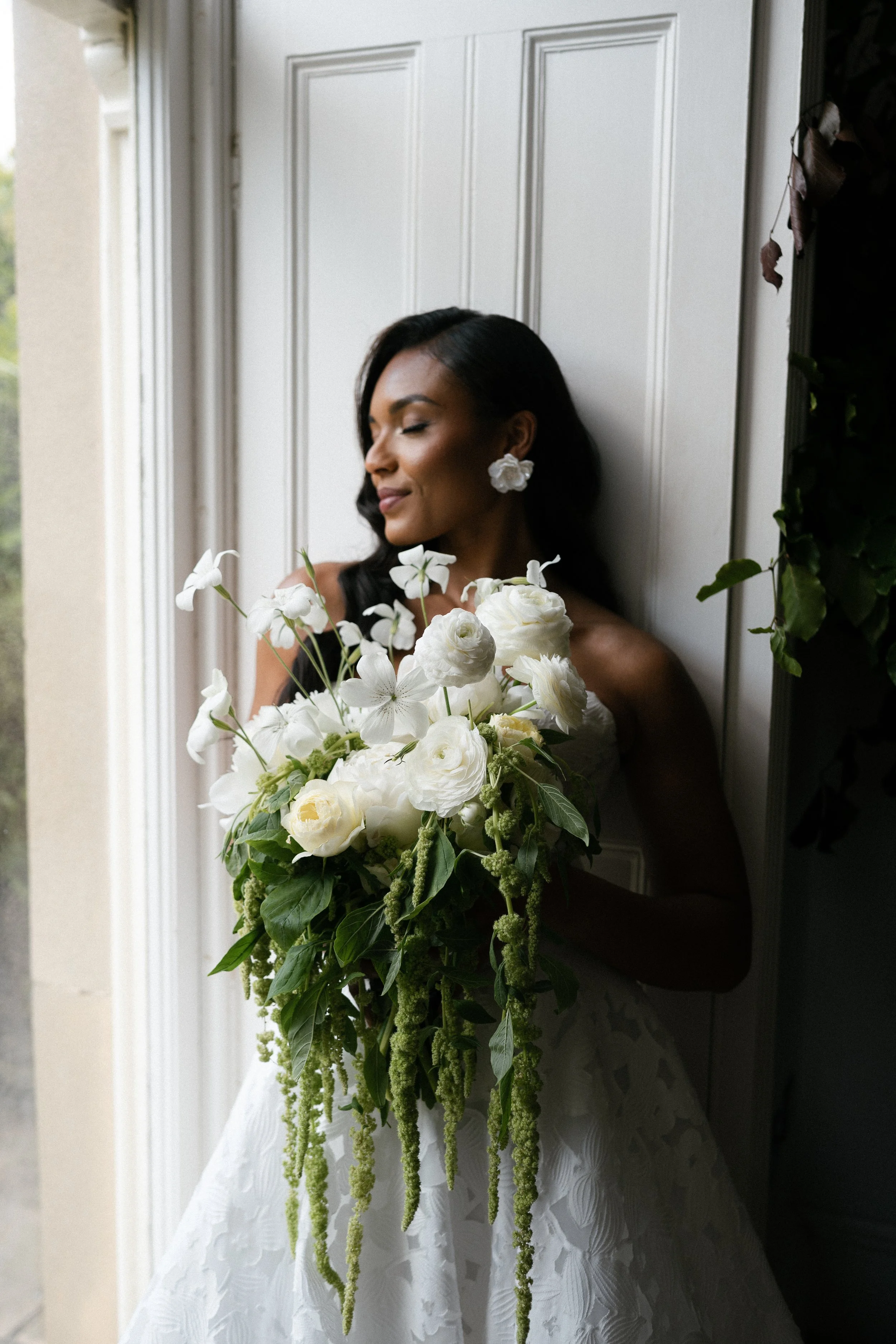 A woman with dark hair and earrings, wearing a white dress, standing indoors near a window, holding a large bouquet of white flowers, with her eyes closed and a content expression.