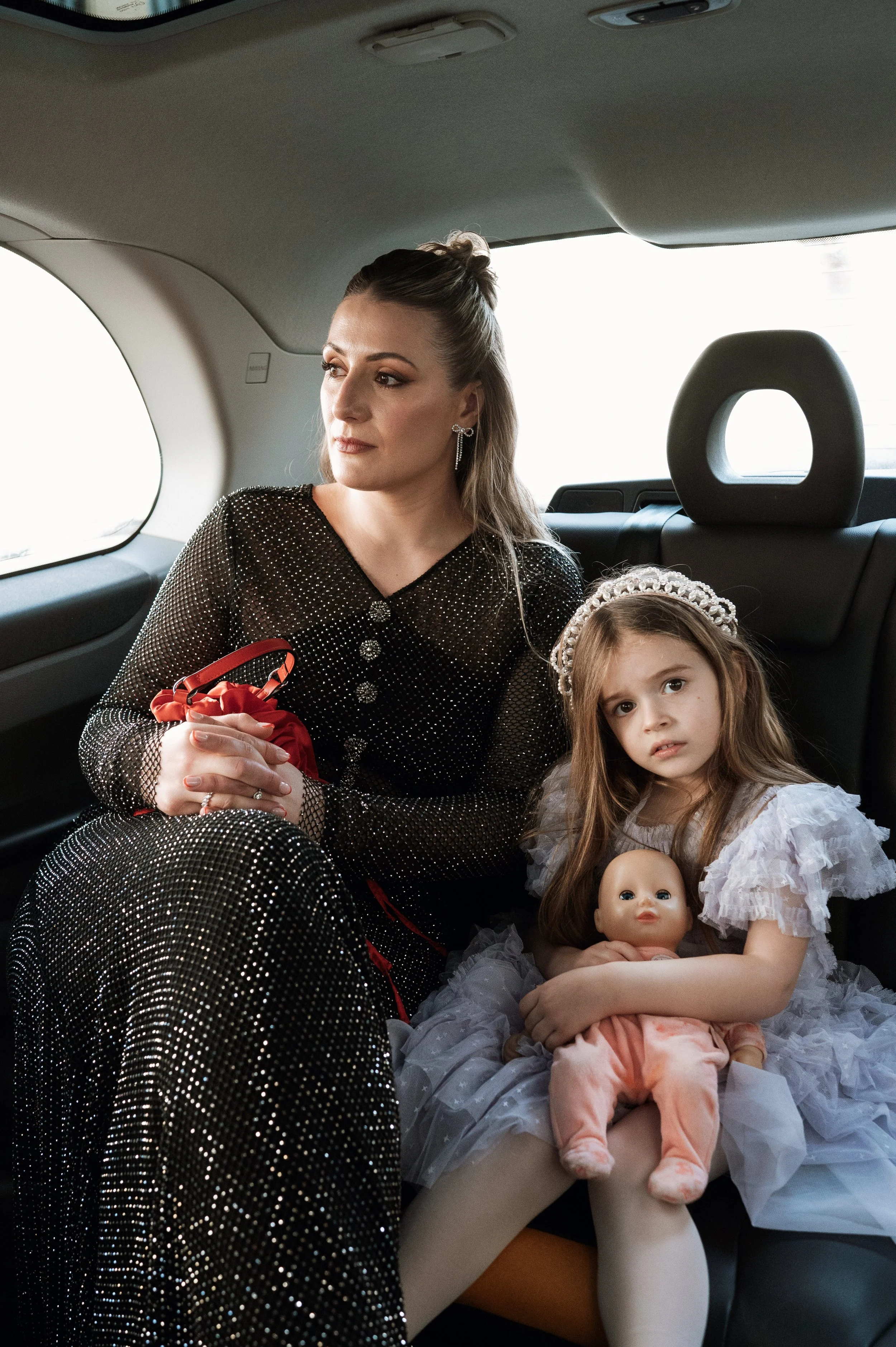 A woman and a young girl sitting in the backseat of a car. The woman has light skin, brown hair styled in an updo, and is wearing a black, sparkly, long-sleeved dress. The girl has light skin, long brown hair, a pearl headband, and a white, ruffled d