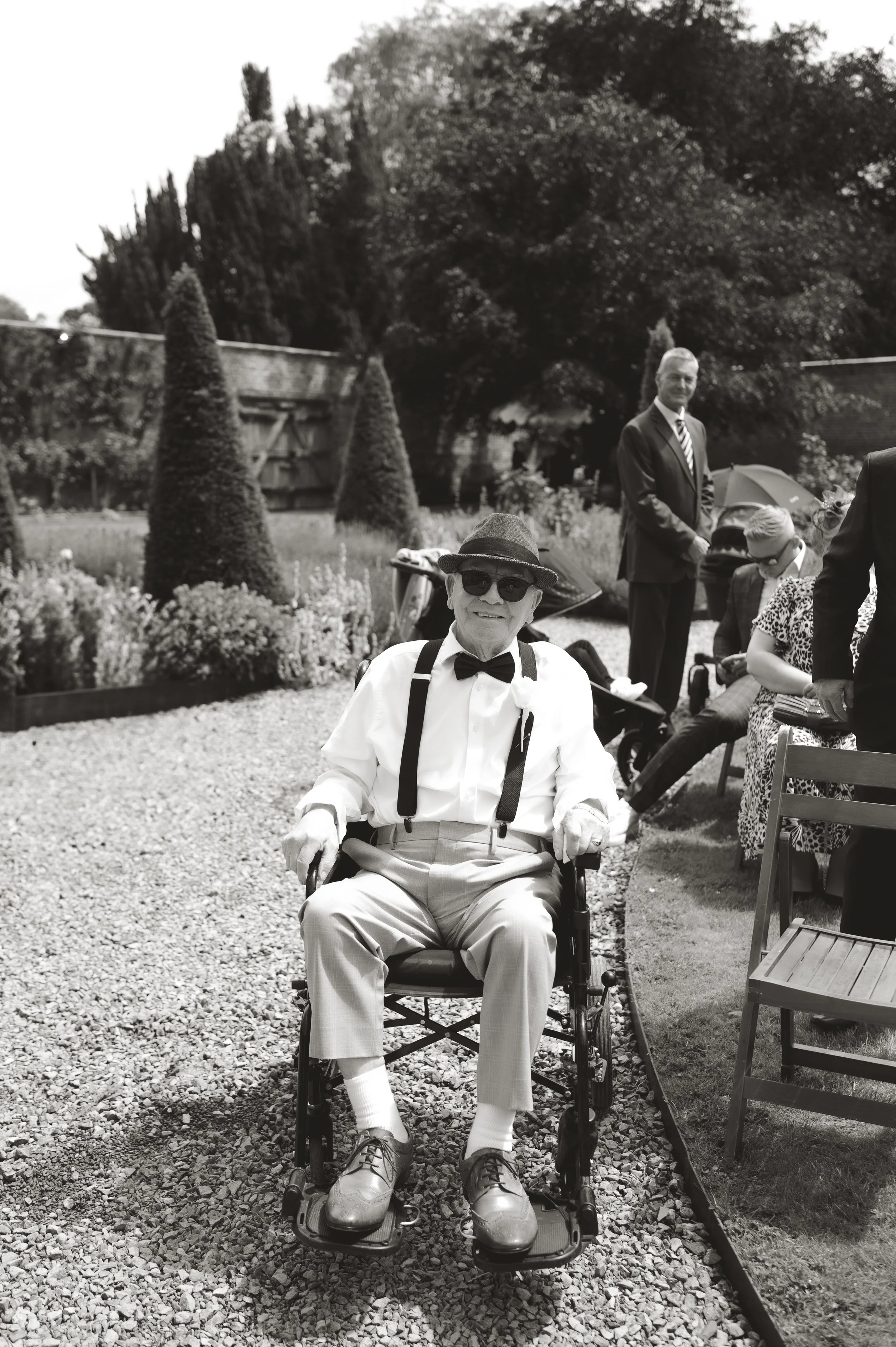 An elderly man dressed in a white shirt, bow tie, suspenders, and light-colored pants, sitting in a wheelchair outdoors at a garden party. He is wearing sunglasses and a fedora hat. In the background, there are guests, some sitting and some standing,