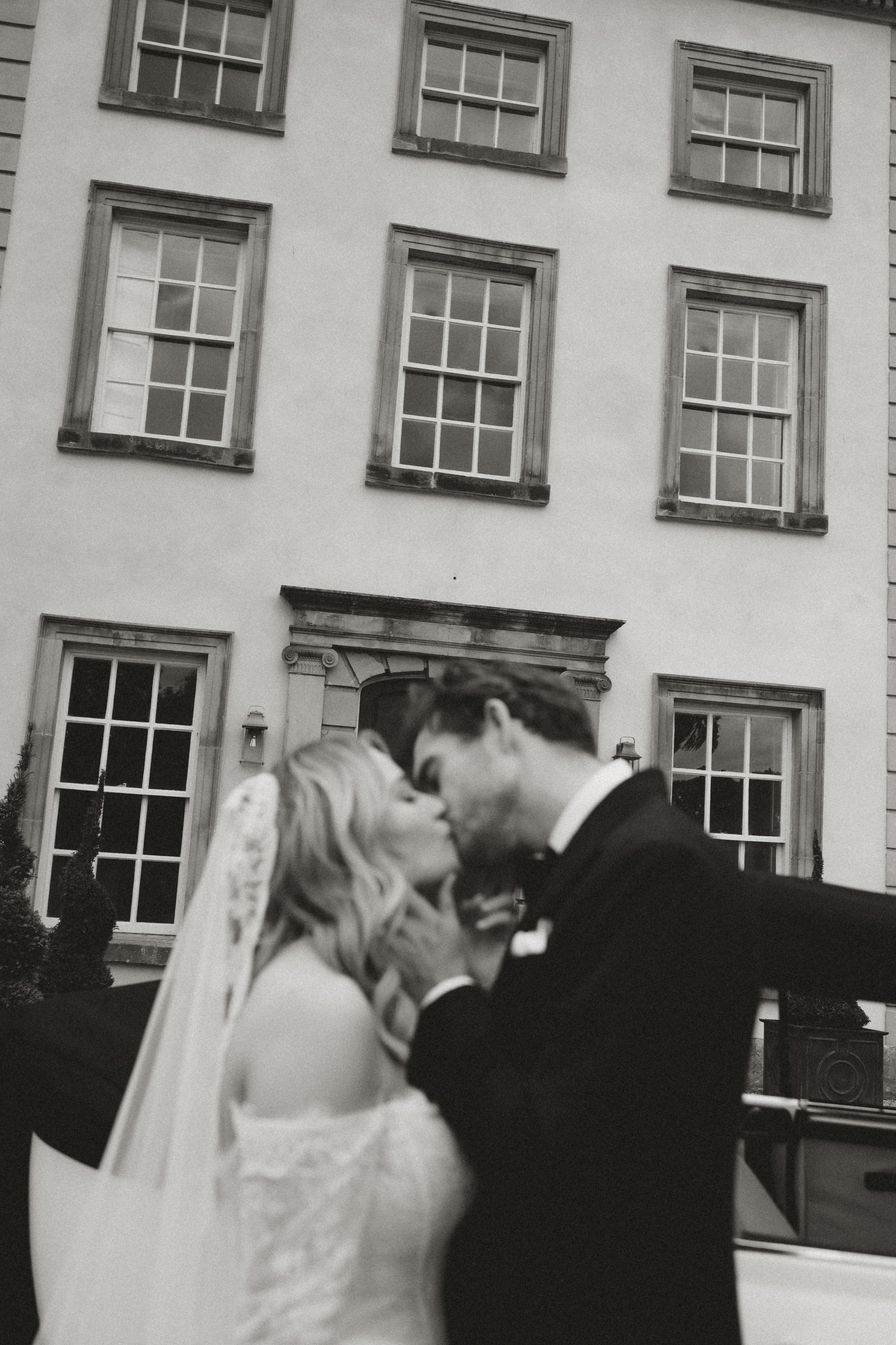 A black and white photo of a bride and groom kissing outside in front of a building with multiple windows.