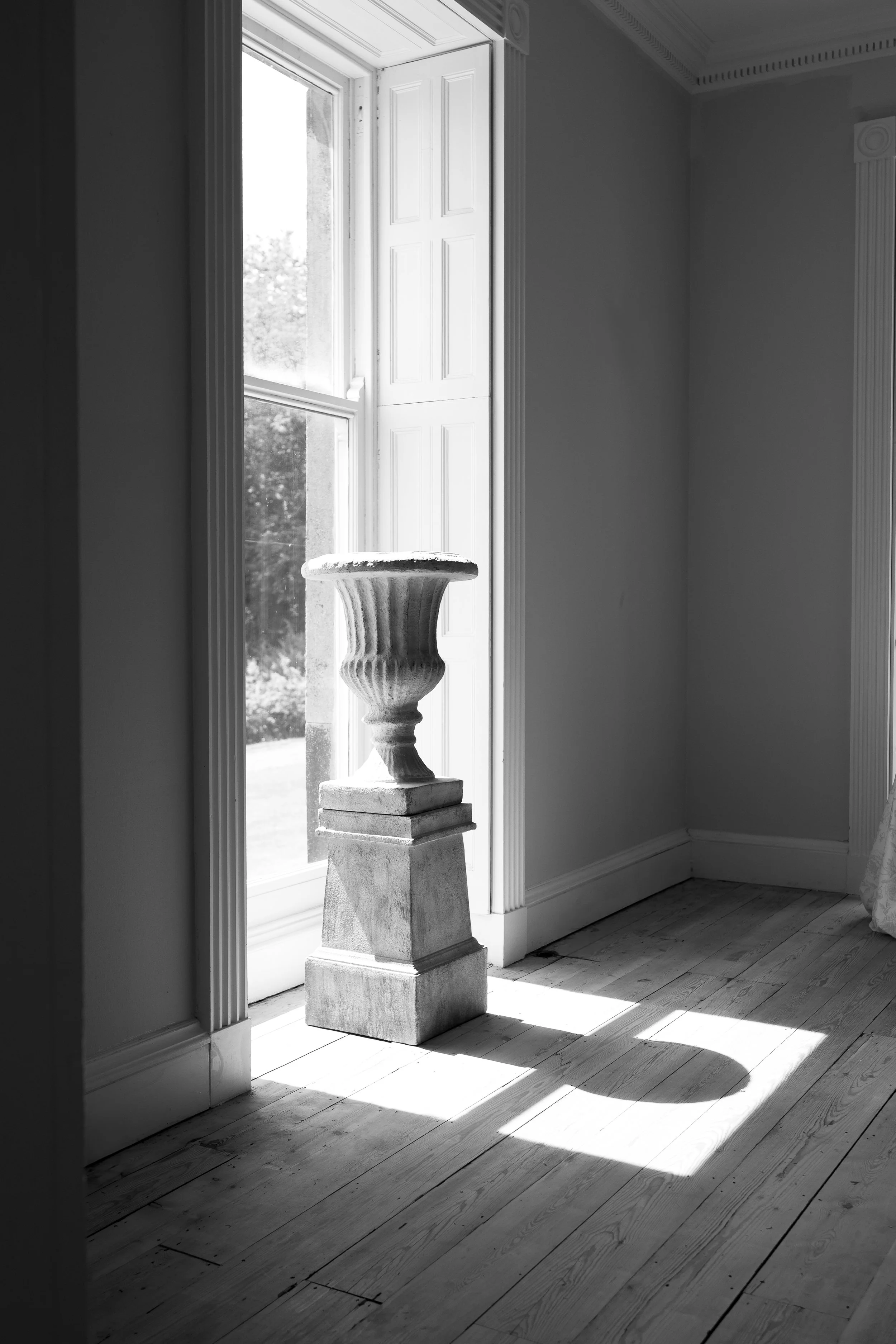A sunlit black and white photo of a stone pedestal with an ornate urn-shaped top standing in front of a tall window with wooden shutters, casting a shadow on the wooden floor.