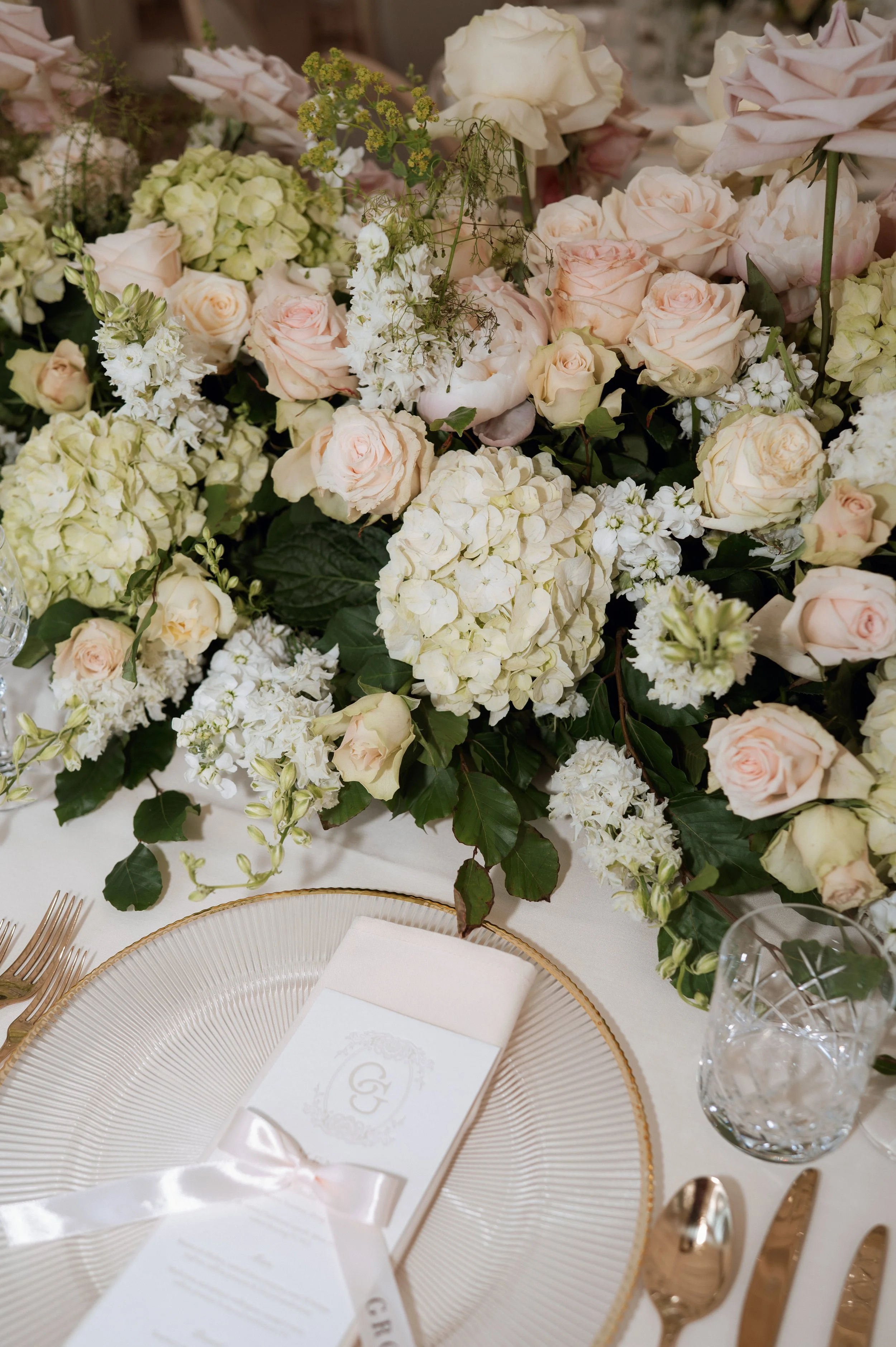 A table setting with a large floral centerpiece of white and pale pink roses, white hydrangeas, and greenery. There is a gold-trimmed clear plate, gold utensils, a crystal glass with ice, and a wrapped menu with a monogrammed initial 'G' and a satin ribbon.