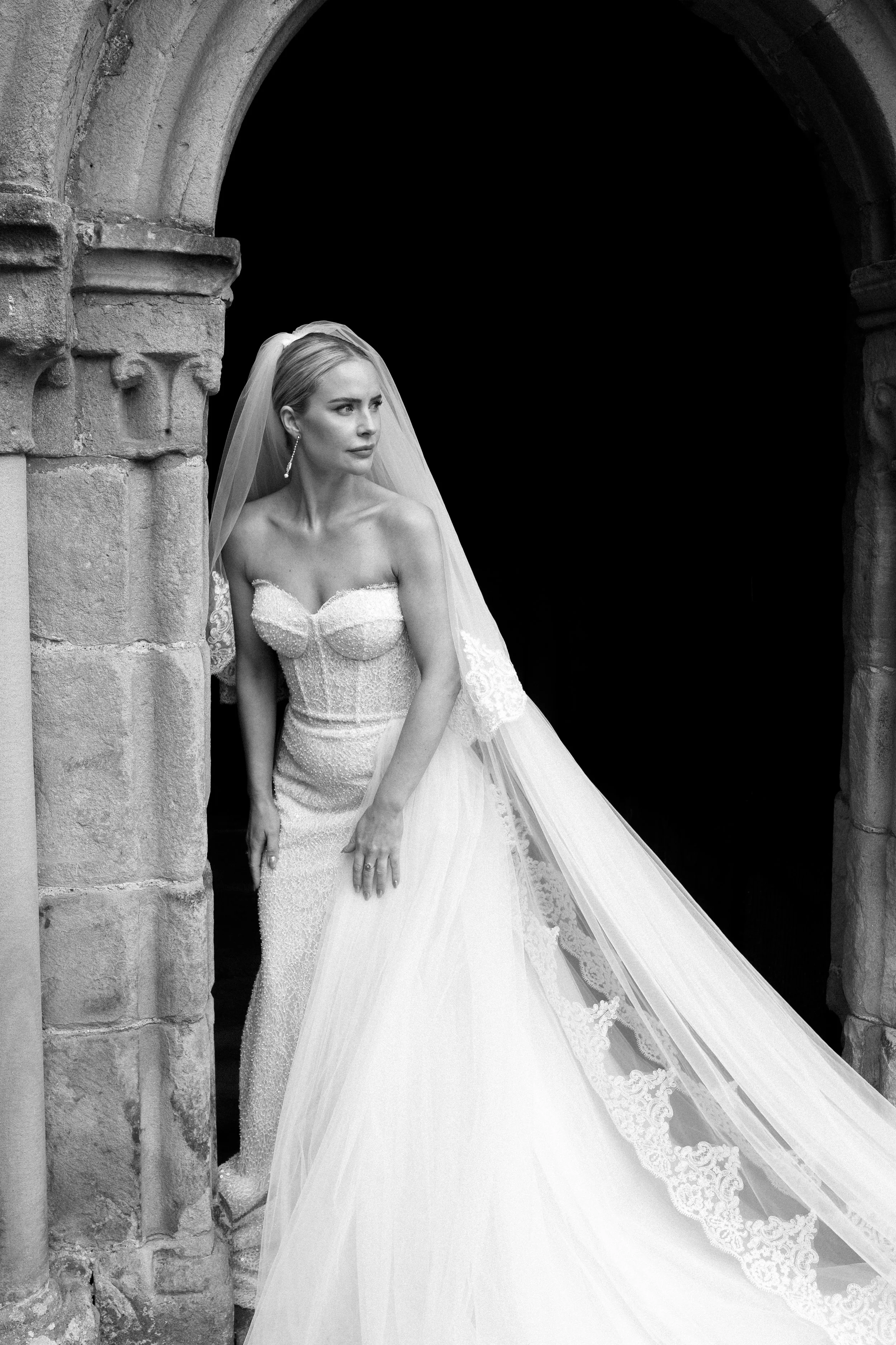 A bride in a wedding gown and veil standing in the doorway of a stone archway, looking to the side with a contemplative expression.