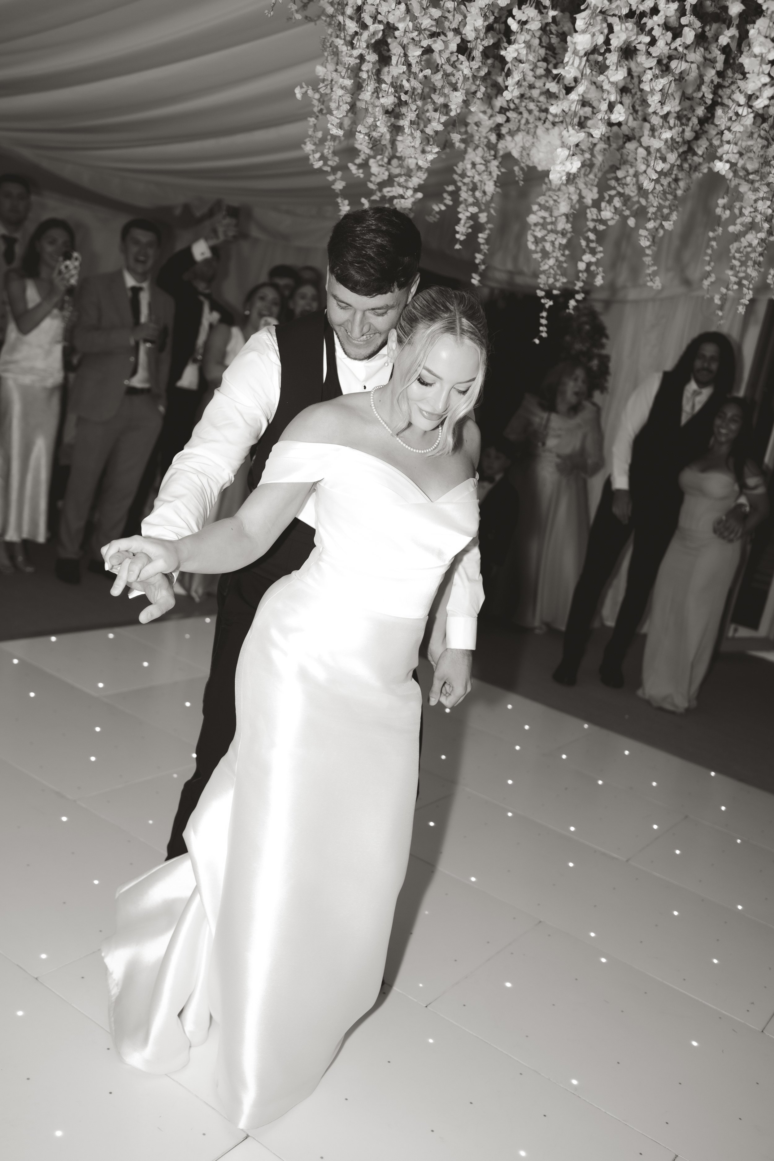 A bride and groom dancing at their wedding reception, surrounded by guests, with a floral ceiling decoration overhead.