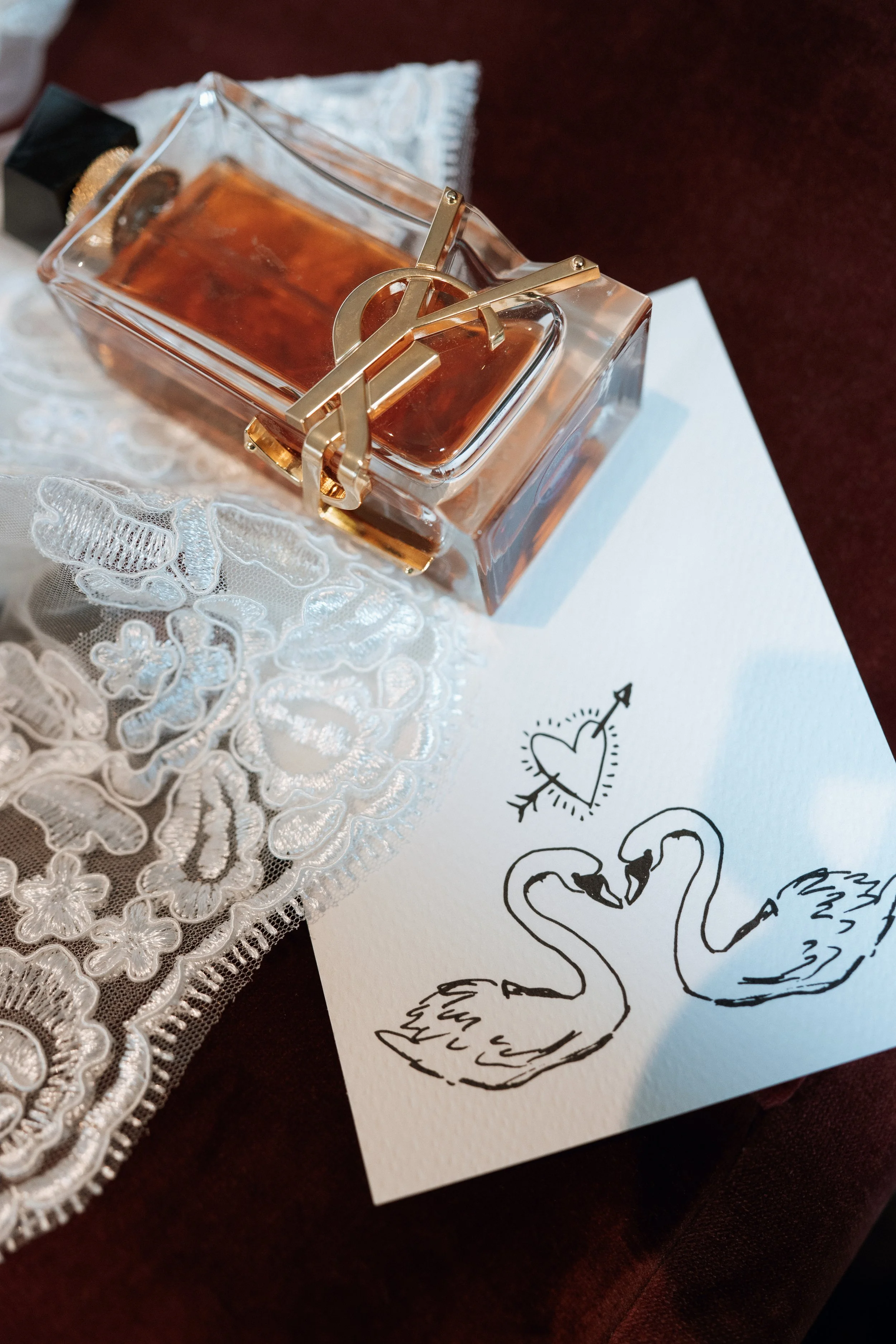 A clear glass perfume bottle with a gold Yves Saint Laurent logo on top, placed on a lace cloth. Next to it is a white card with a black line drawing of two swans facing each other, forming a heart shape with their necks, and a small heart with an ar