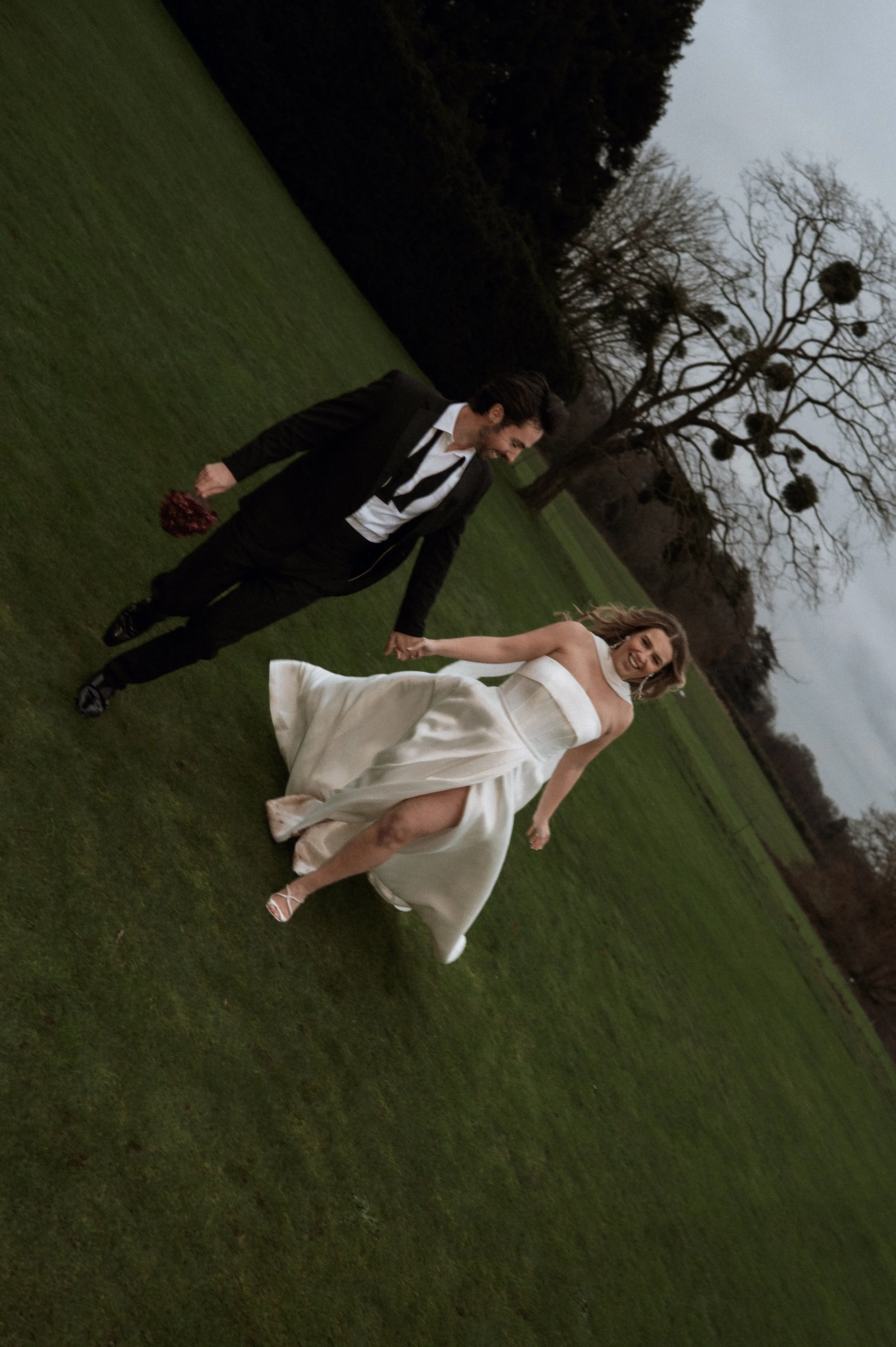 A joyful woman in a white wedding dress and a man in a black suit walk hand in hand across a green field, with trees and cloudy sky in the background.