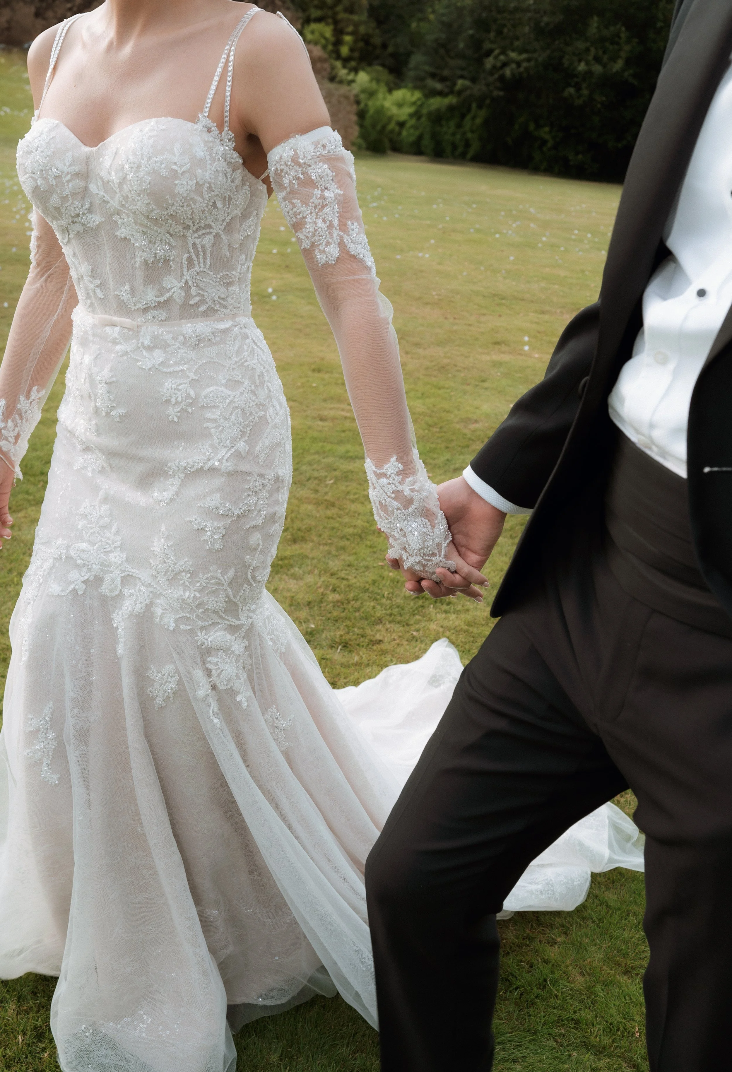 A bride in a white lace wedding dress holding hands with a man in a black tuxedo outdoors on grass.