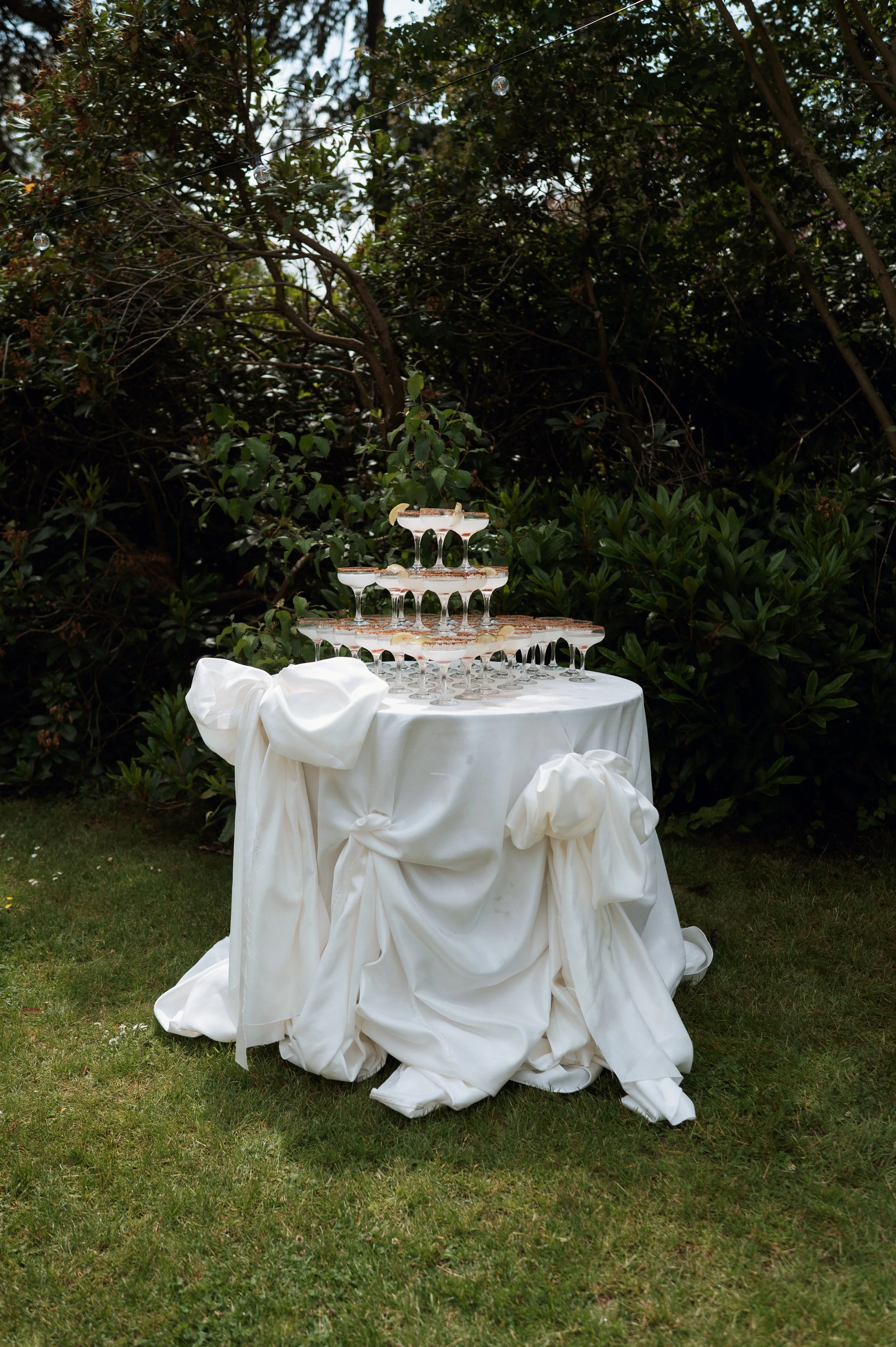 A tiered champagne glass pyramid on a white draped table outdoors, surrounded by greenery.