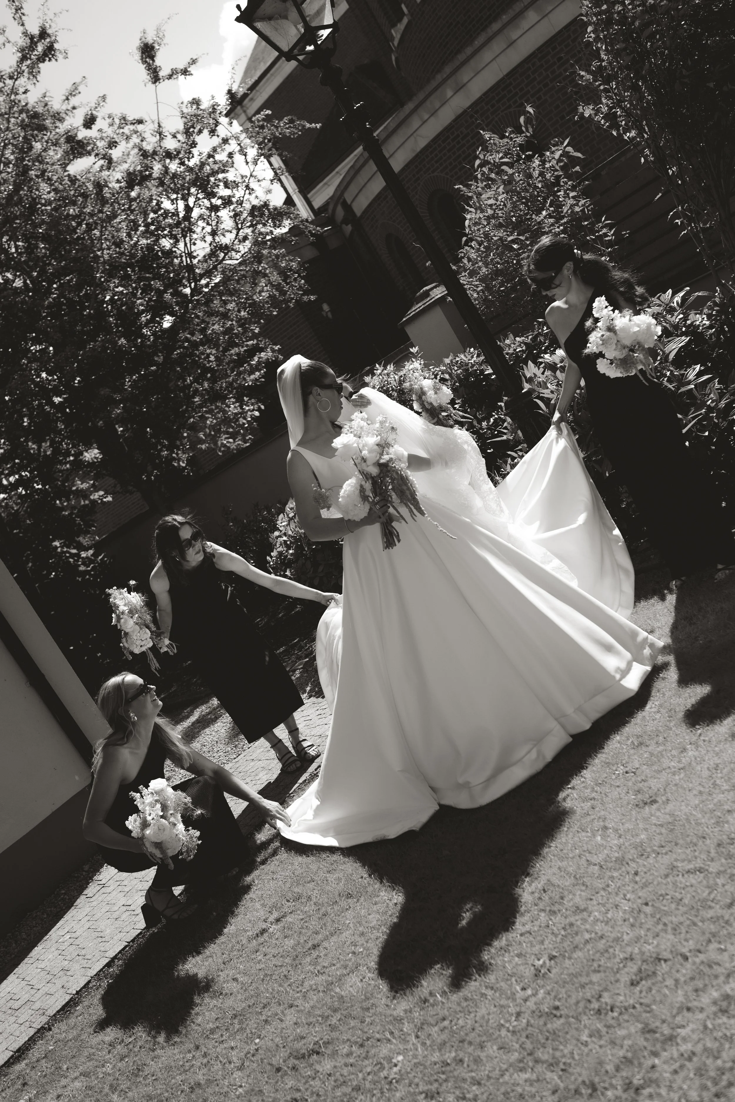 A bride in a wedding dress holding a bouquet, surrounded by four bridesmaids holding bouquets, outdoors on a sunny day.