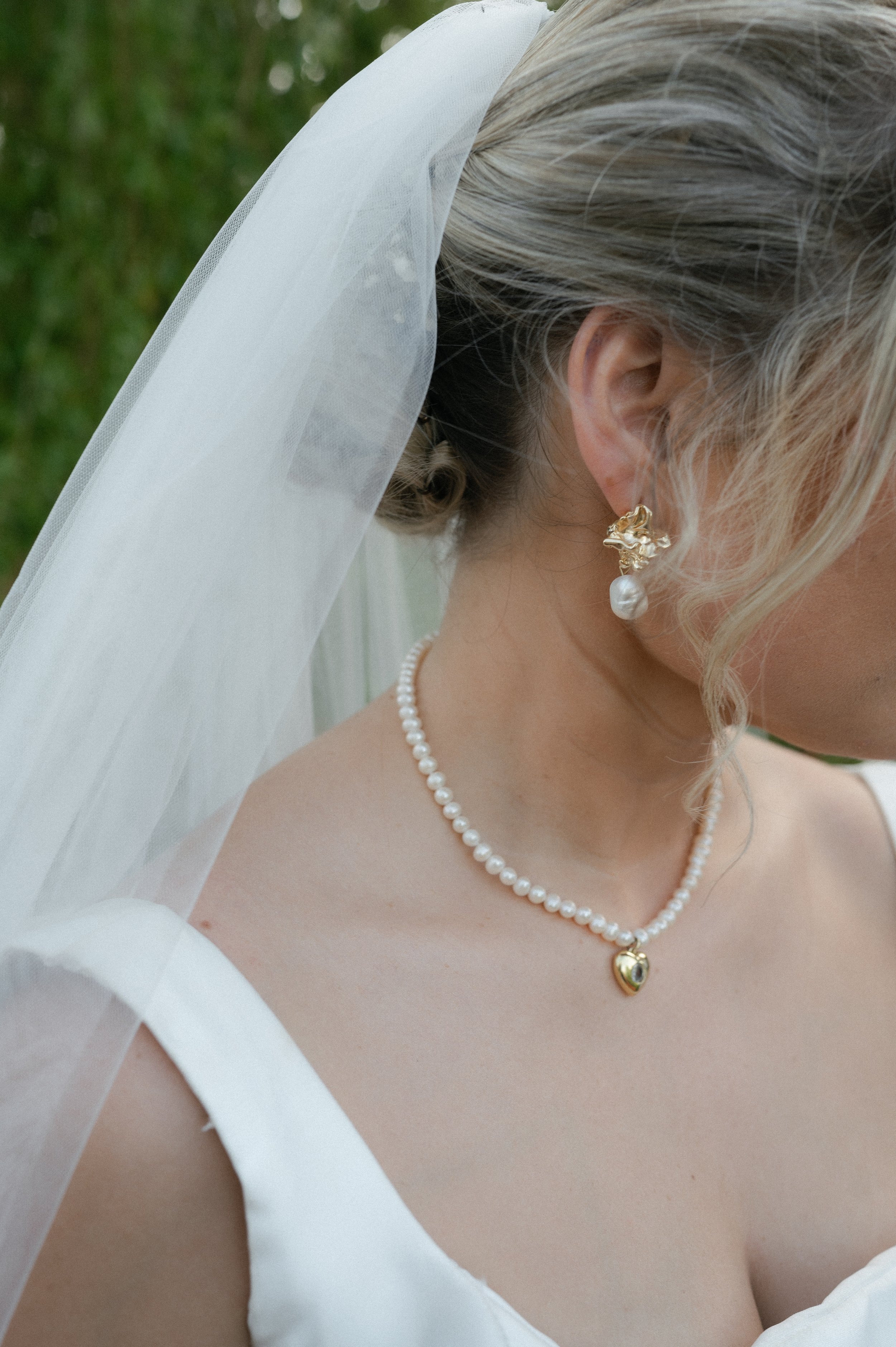 Close-up of a bride wearing pearl jewelry, a pearl necklace with a gold heart pendant, and pearl earrings, with a white veil and a white dress, outdoors with blurred greenery in the background.
