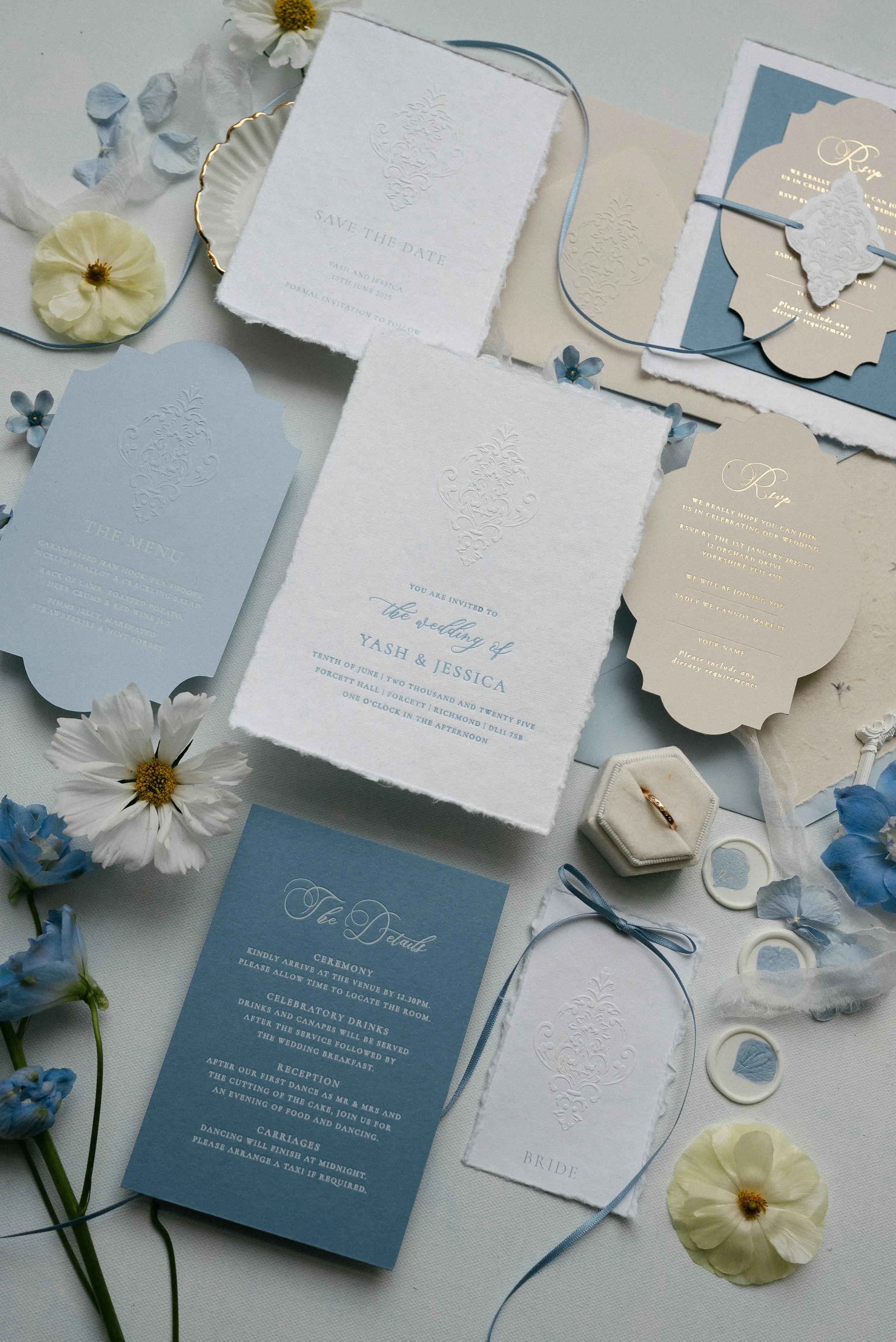Flat lay of wedding stationery and accessories including invitations, menu, save-the-date card, RSVP card, wedding details card, ring box with rings, flower petals, and flowers in shades of white and blue.