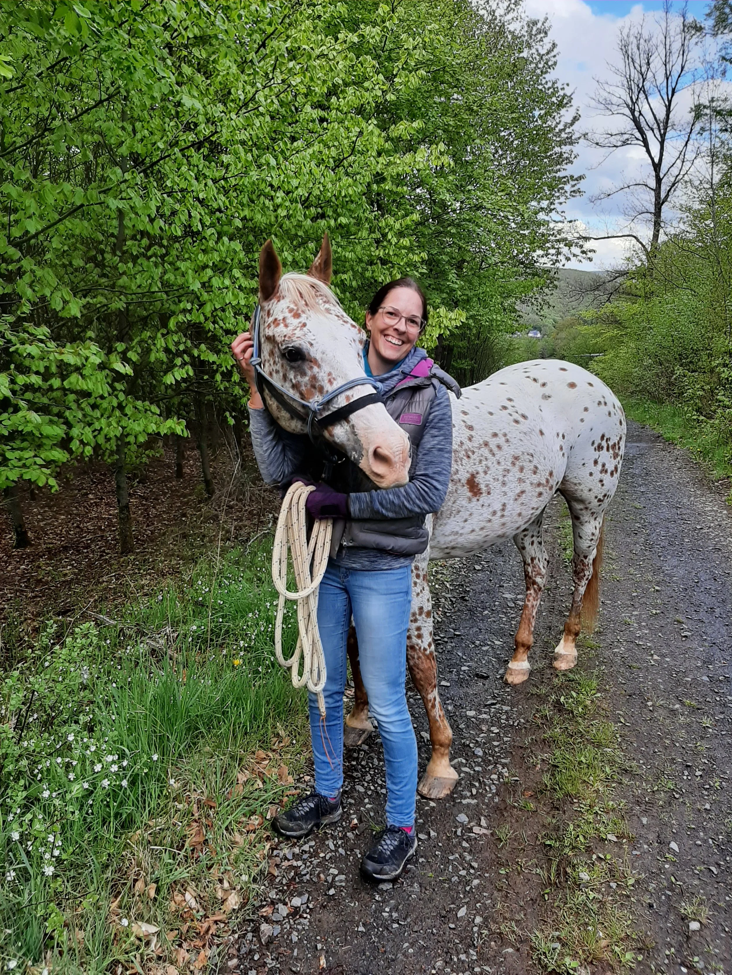 Frau mit einem Pferd auf einem Waldweg, grüne Bäume im Hintergrund.