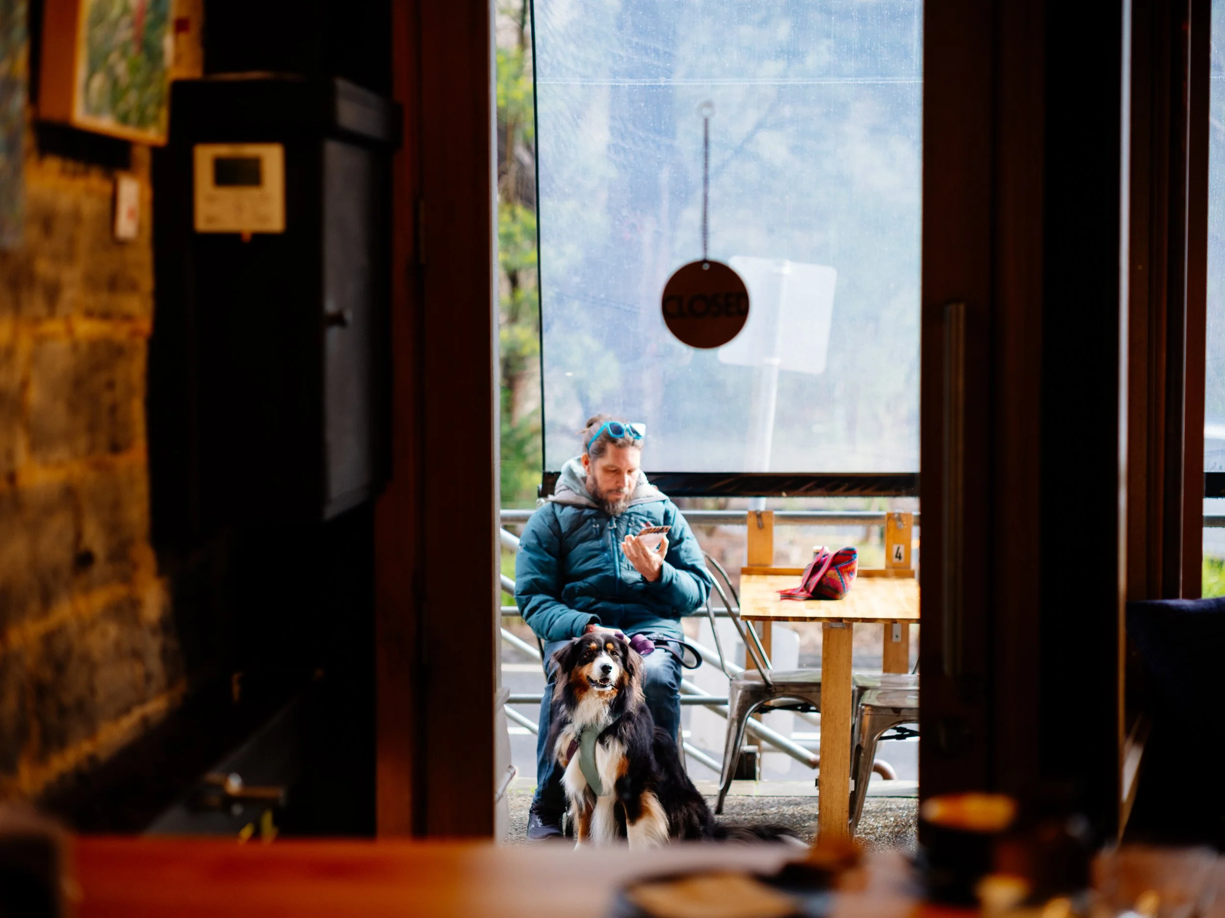 A person sitting with a dog outside, viewed through an open doorway with a "Closed" sign hanging in the door window. The person is wearing sunglasses on their head and a blue jacket, and the dog is an Australian Shepherd.