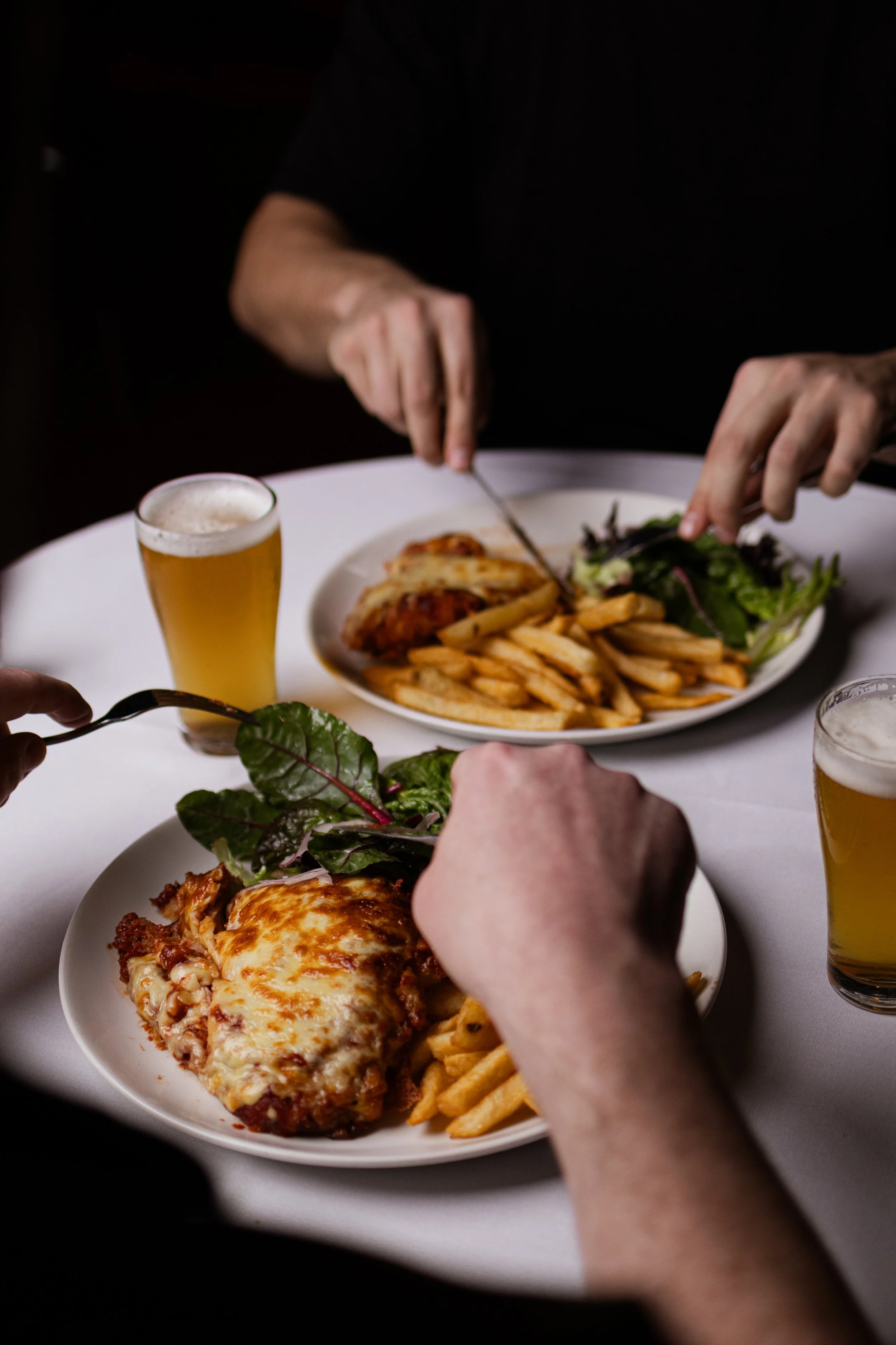 People eating at a white table with plates of lasagna, fries, salads, and glasses of beer.