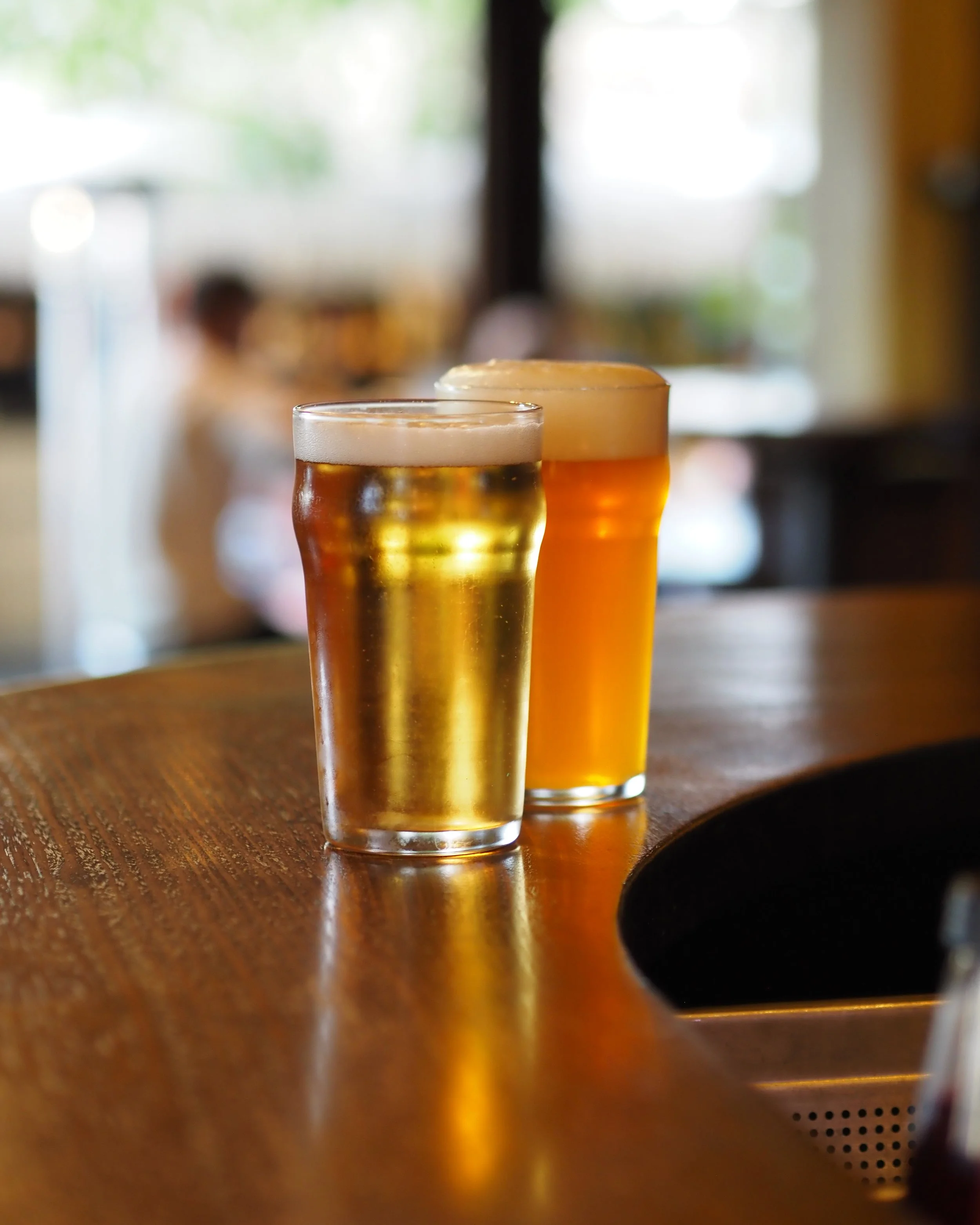 Two glasses of beer, one pale and one darker, on a wooden bar counter inside a bar or pub.