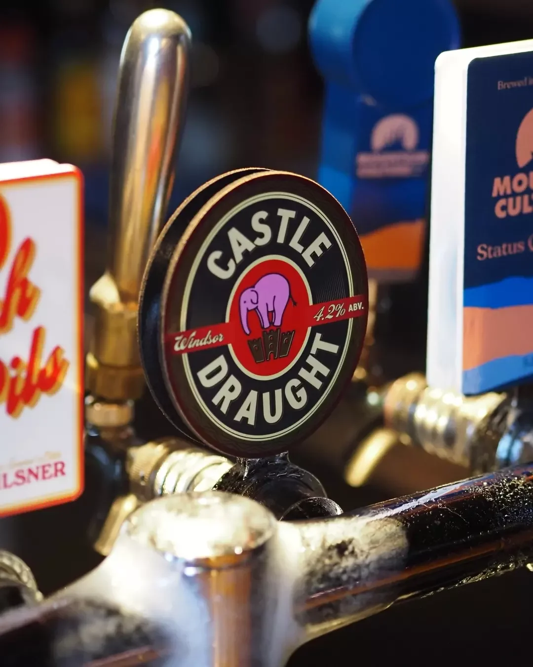 Close-up of a Castle Draught beer tap with an elephant logo, surrounded by other beer taps at a bar.