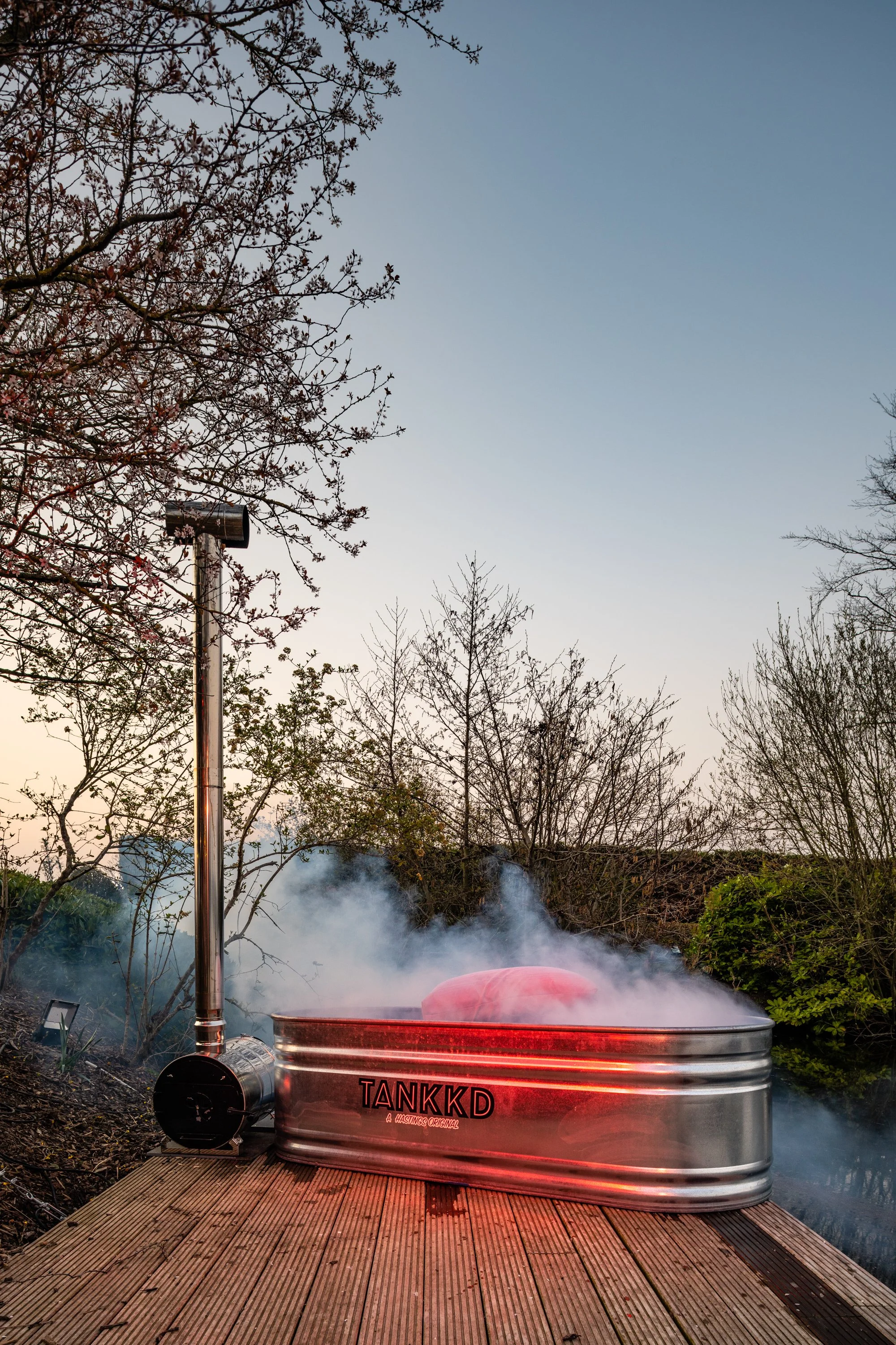 A metal hot tub labeled 'TANKKD' emitting steam on a wooden deck outdoors, with trees and a clear sky in the background.