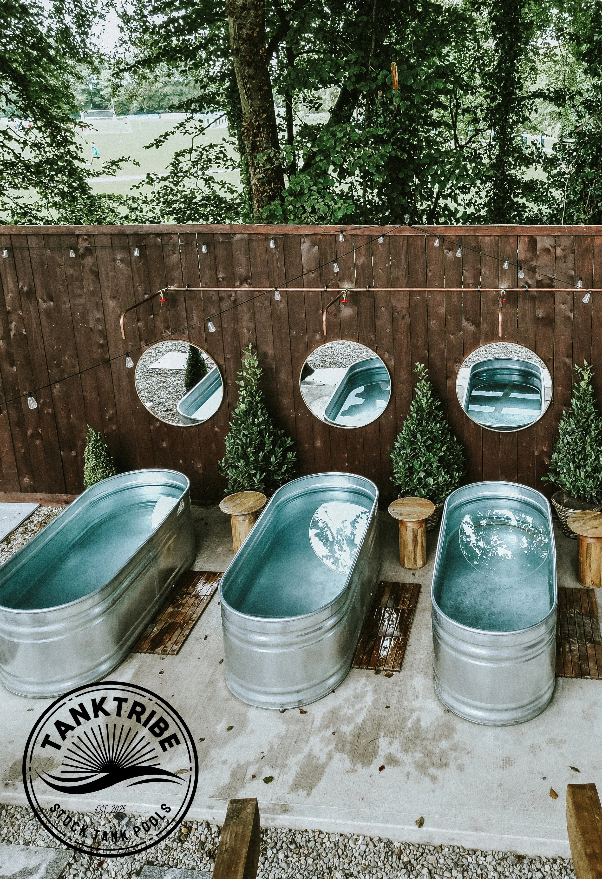 Three metal tubs filled with water, placed on a concrete surface, with small wooden stools beside them. A wooden fence with three round mirrors and small green plants in pots is in the background. String lights are hanging above.