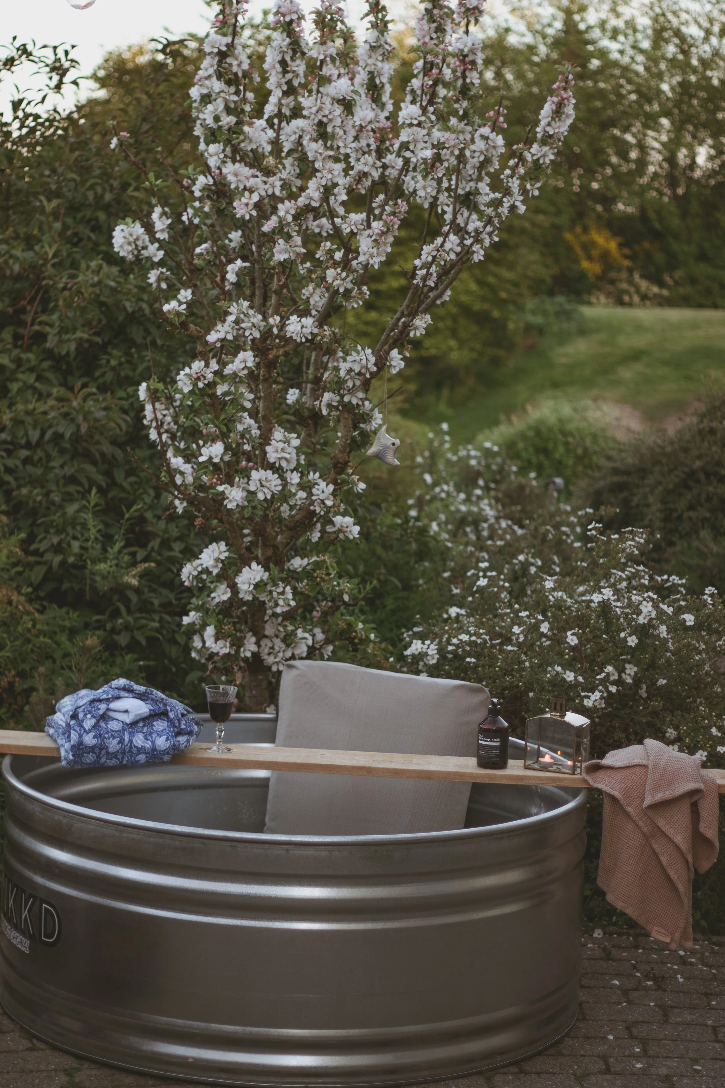 An outdoor bathing setup with a Stock Tank, a pillow, a glass of wine, and cozy towels, surrounded by blooming cherry blossom trees.