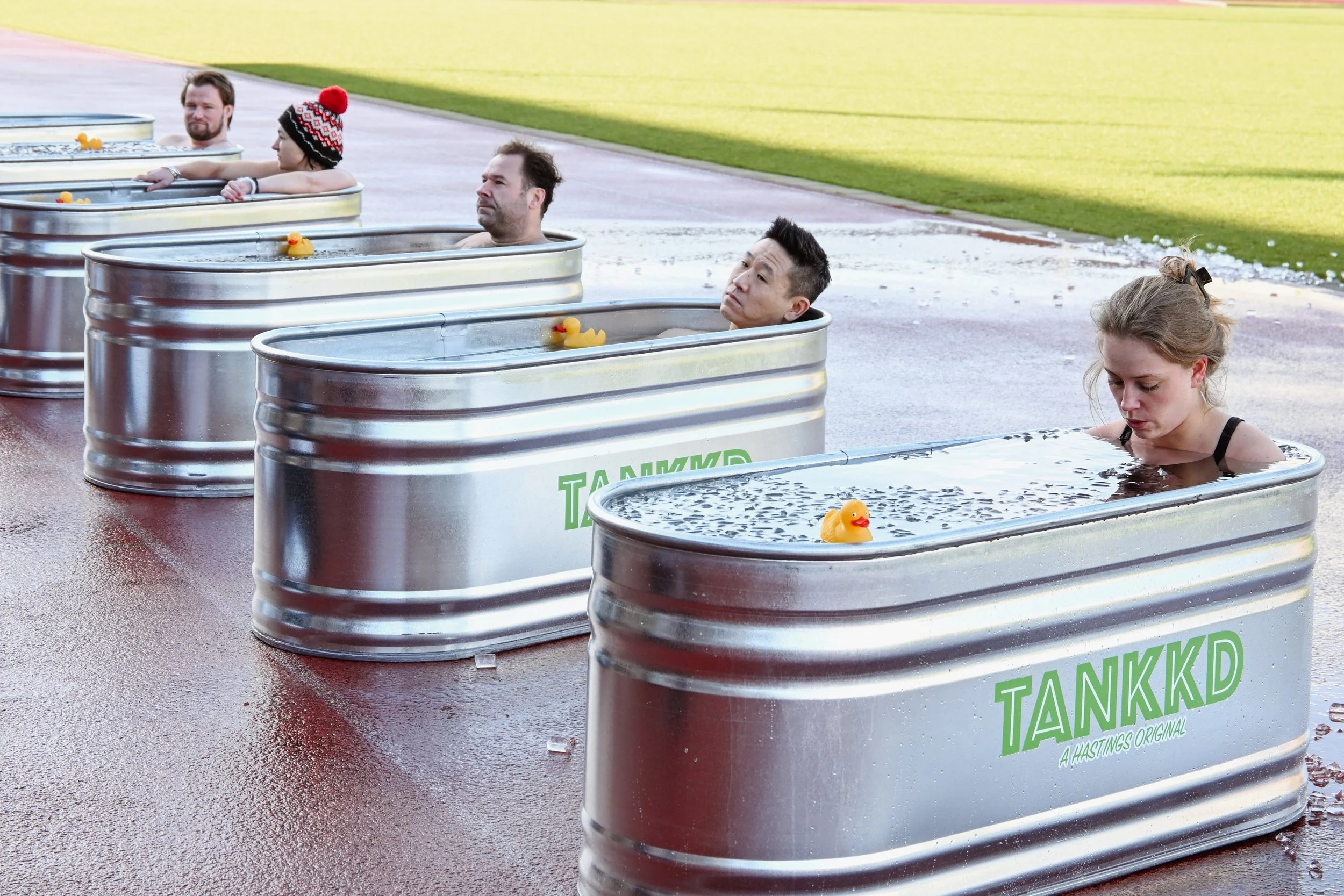 People enjoy a relaxing soak in outdoor tubs filled with water, each tub with a rubber ducky float, in a park setting.