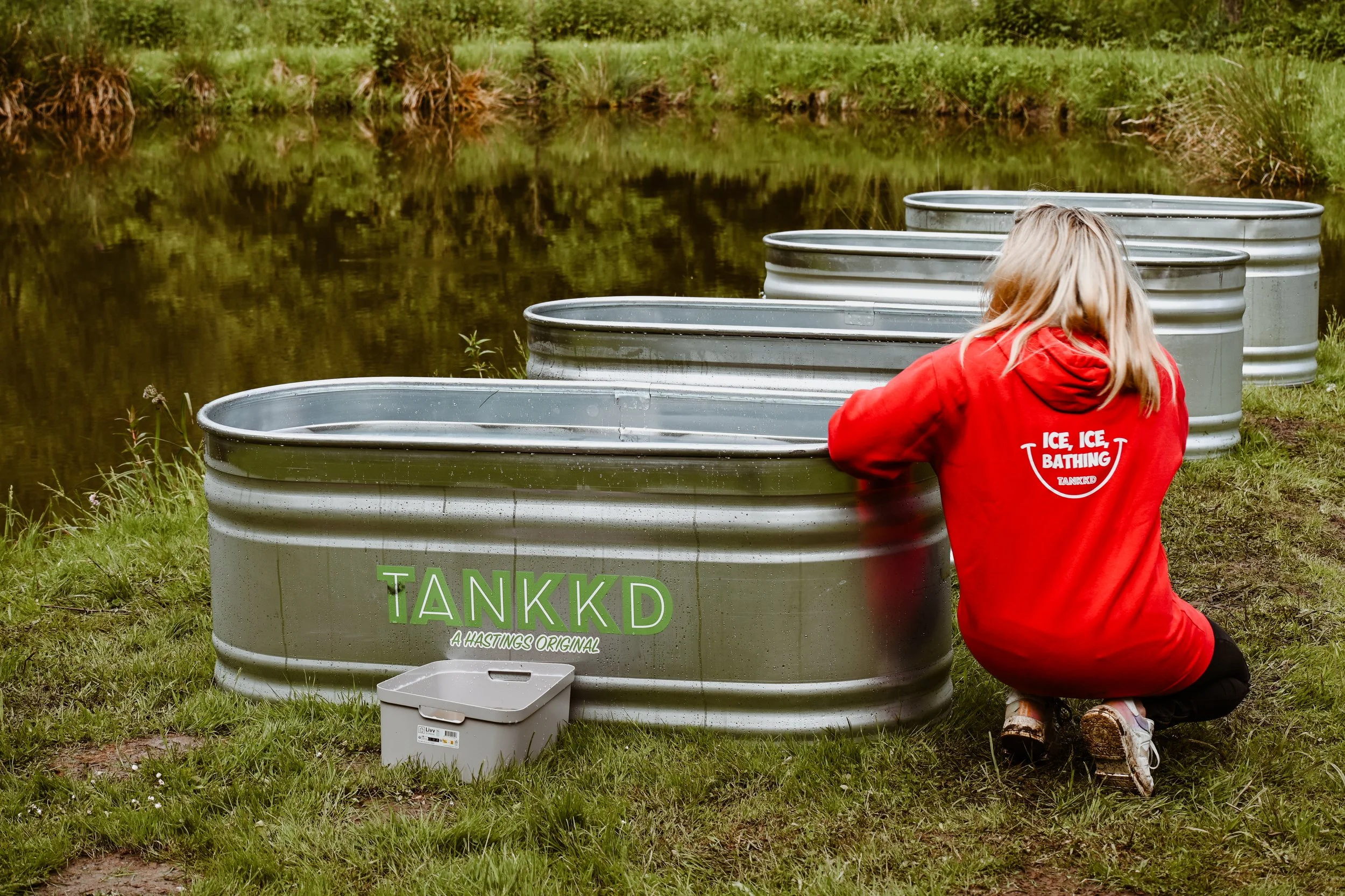A woman in a red hoodie kneels beside a series of large metallic tubs near a pond, with grass and trees in the background.