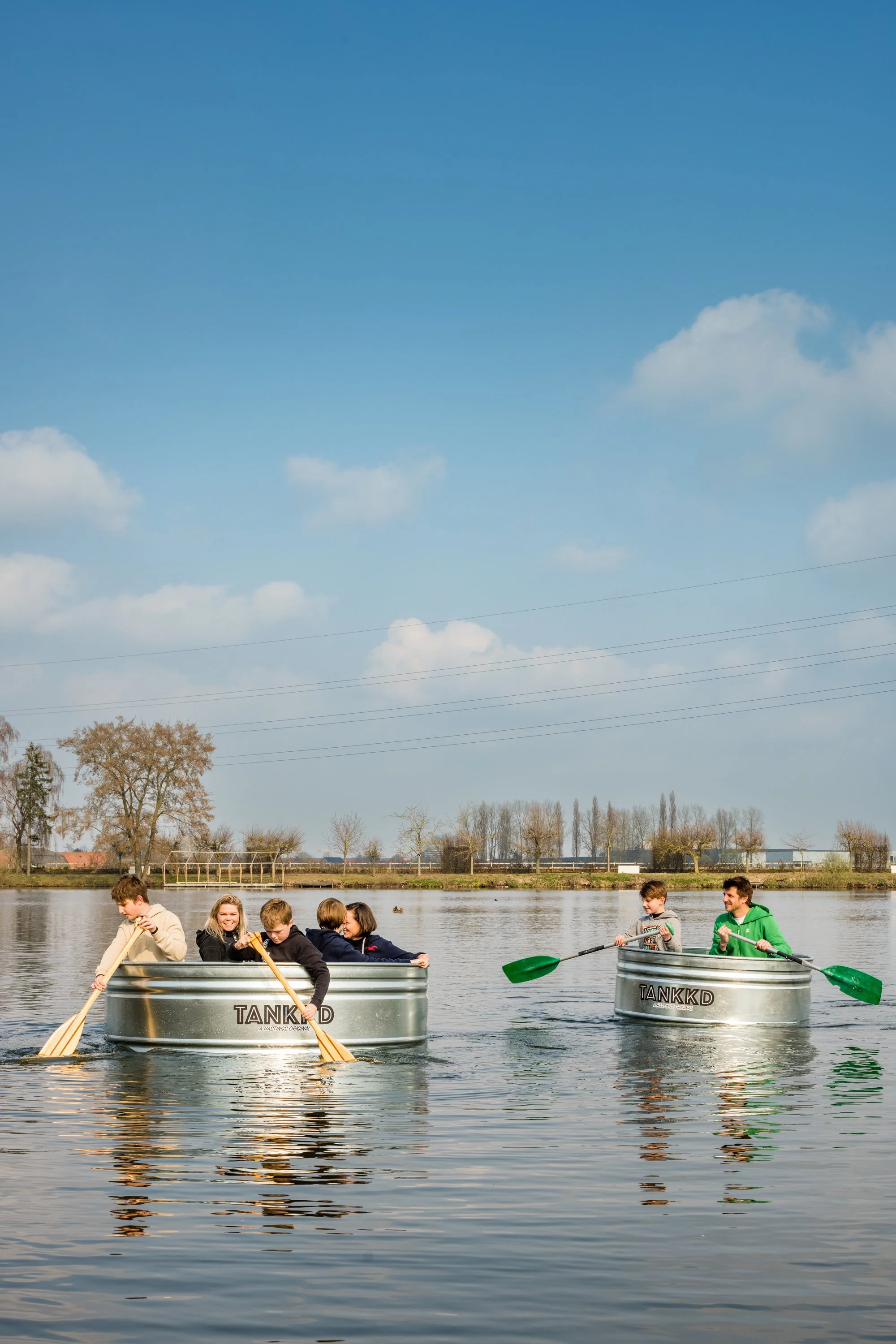Children and an adult in two metal tubs paddling on a calm lake with trees and power lines in the background on a sunny day.