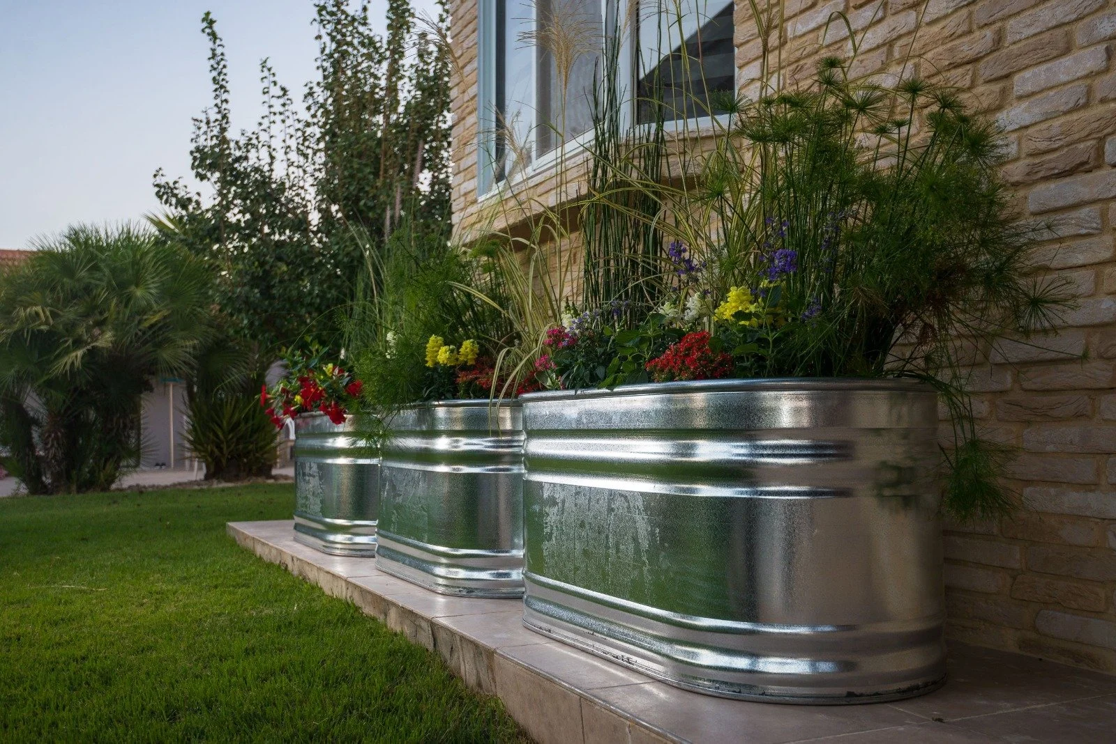 Three large metal planters filled with colorful flowers and green plants, placed on a concrete platform against a stone wall, with a grassy lawn and trees in the background.