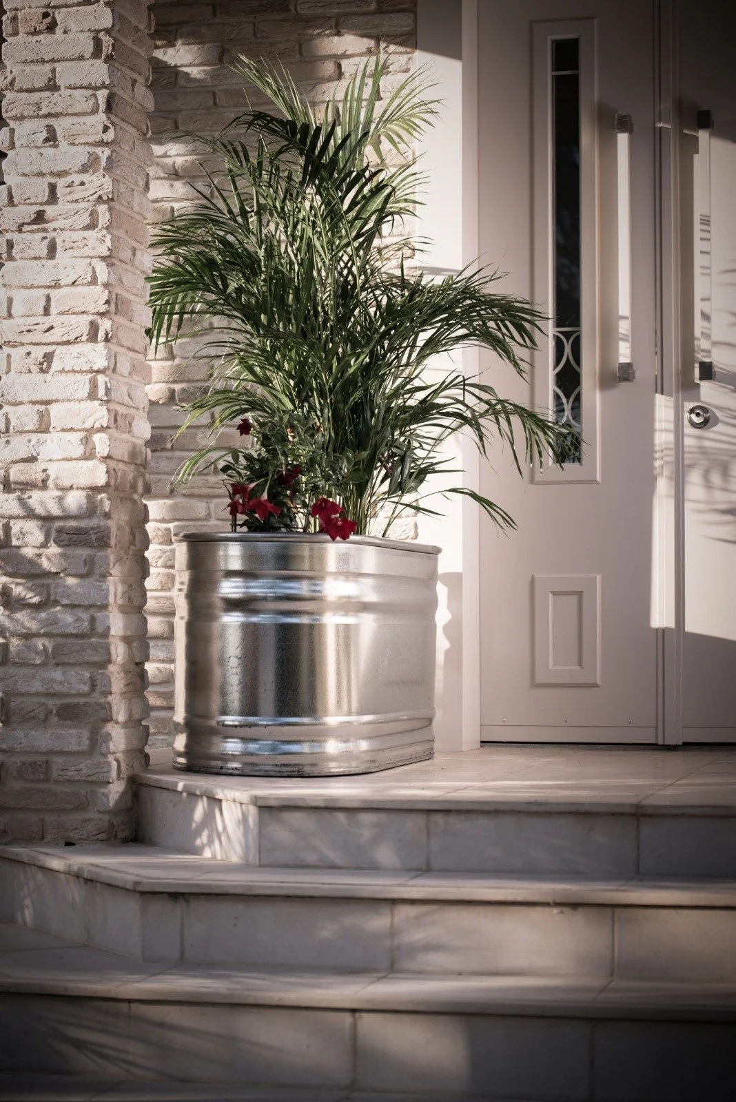 Indoor entryway with a white brick wall, white door, and a large green potted plant in a shiny metallic container on marble steps.