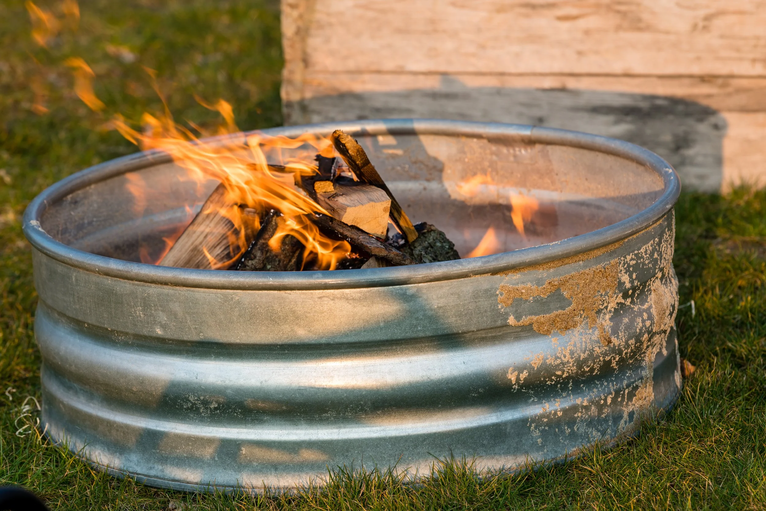 Fire burning in a metal container outdoors on grass with a wooden background.