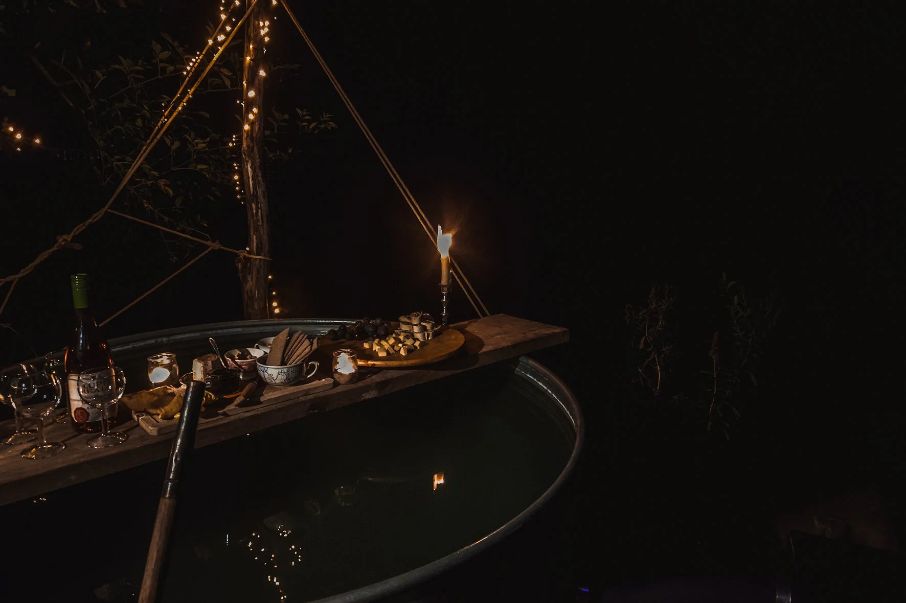 A nighttime outdoor scene featuring a wooden tray with food, drinks, and a lit candle set across a bathtub filled with water. String lights and ropes decorate the background.