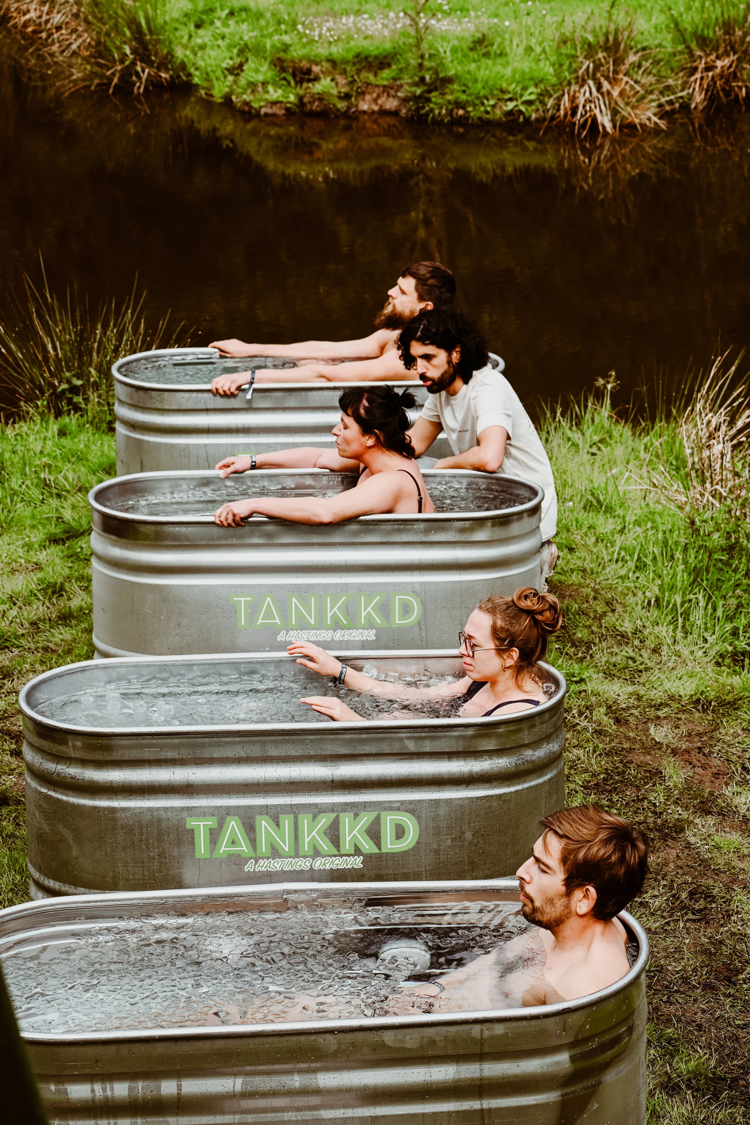 Five people relaxing in ice baths filled with water positioned outdoors near a pond or lake on a grassy bank.