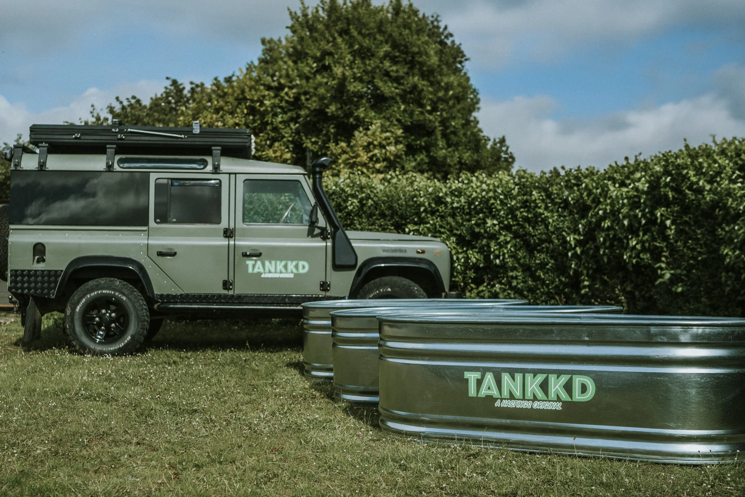 A Landrover defender 110 with the label TANKKD parked next to four large stock tanks with the same label, set on grassy ground with bushes and trees in the background.