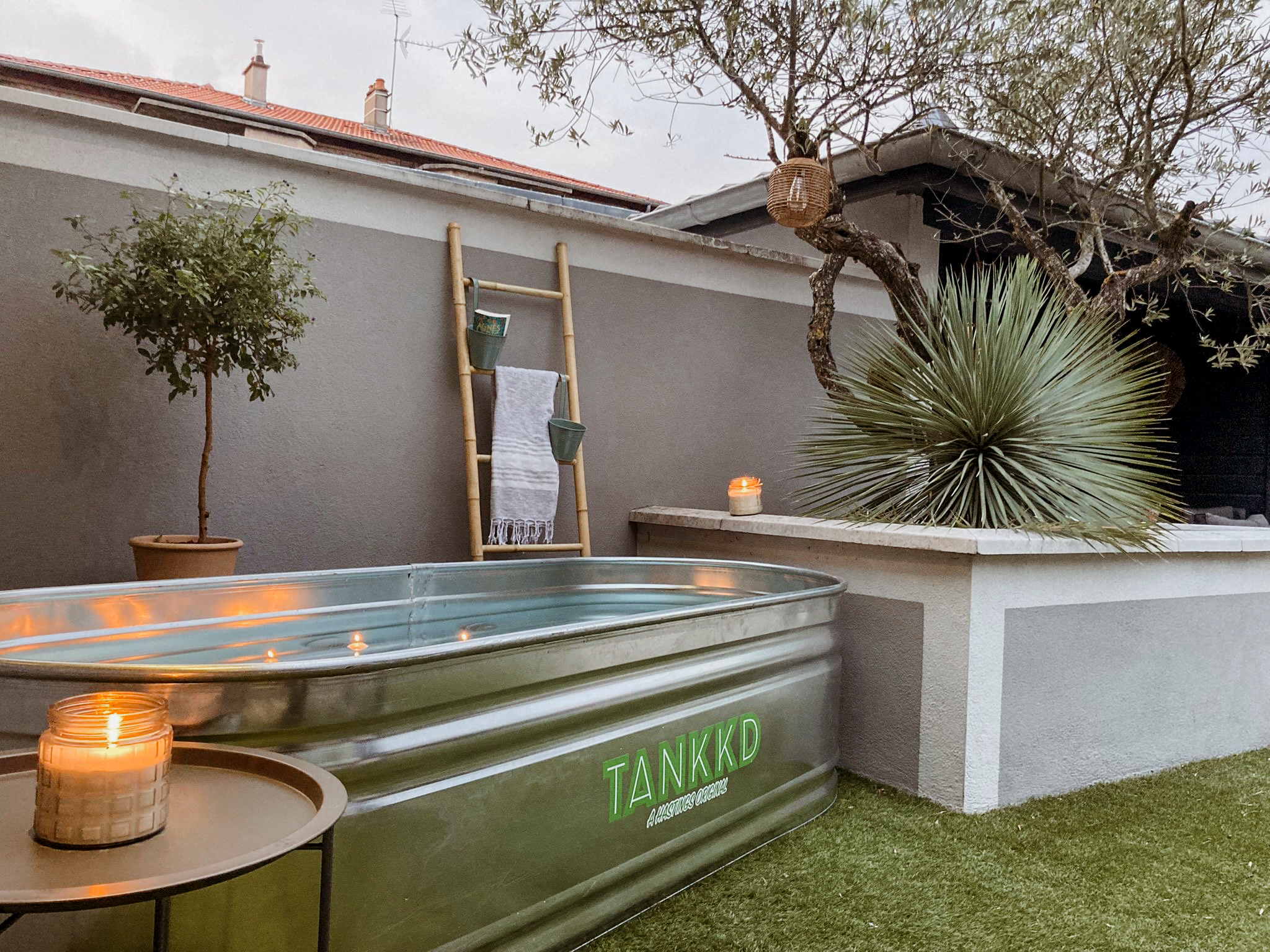 Outdoor backyard scene with a hot tub, lit candles, a small potted tree, a bamboo towel rack, a large spiky plant, and a tree with hanging basket. The scene appears to be during dusk or evening.