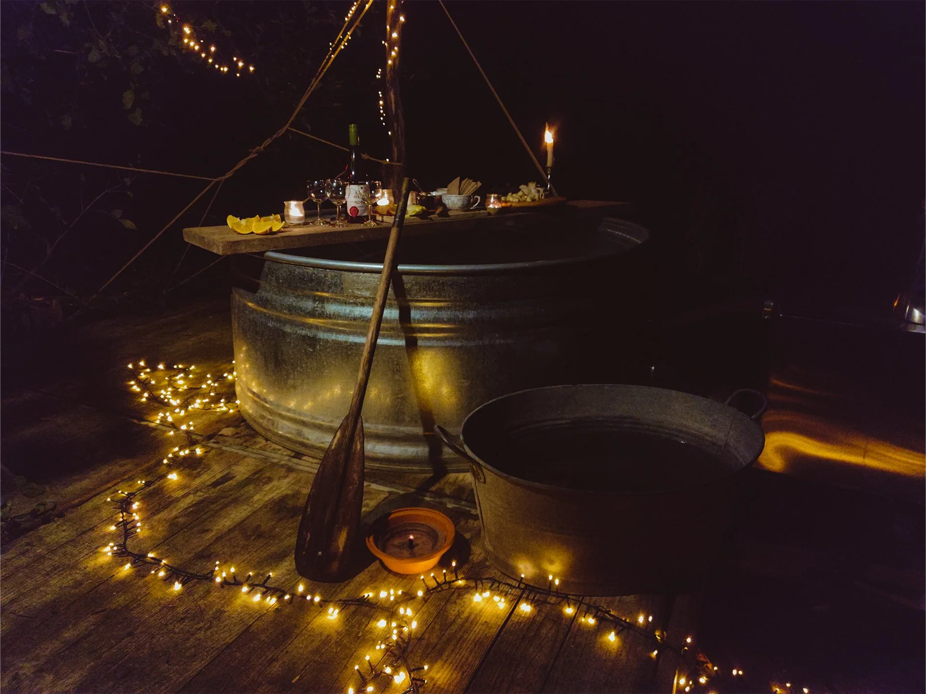 Outdoor romantic setup with a hot tub, string lights, a paddle, candles, wine glasses, and snacks on a wooden deck at night.