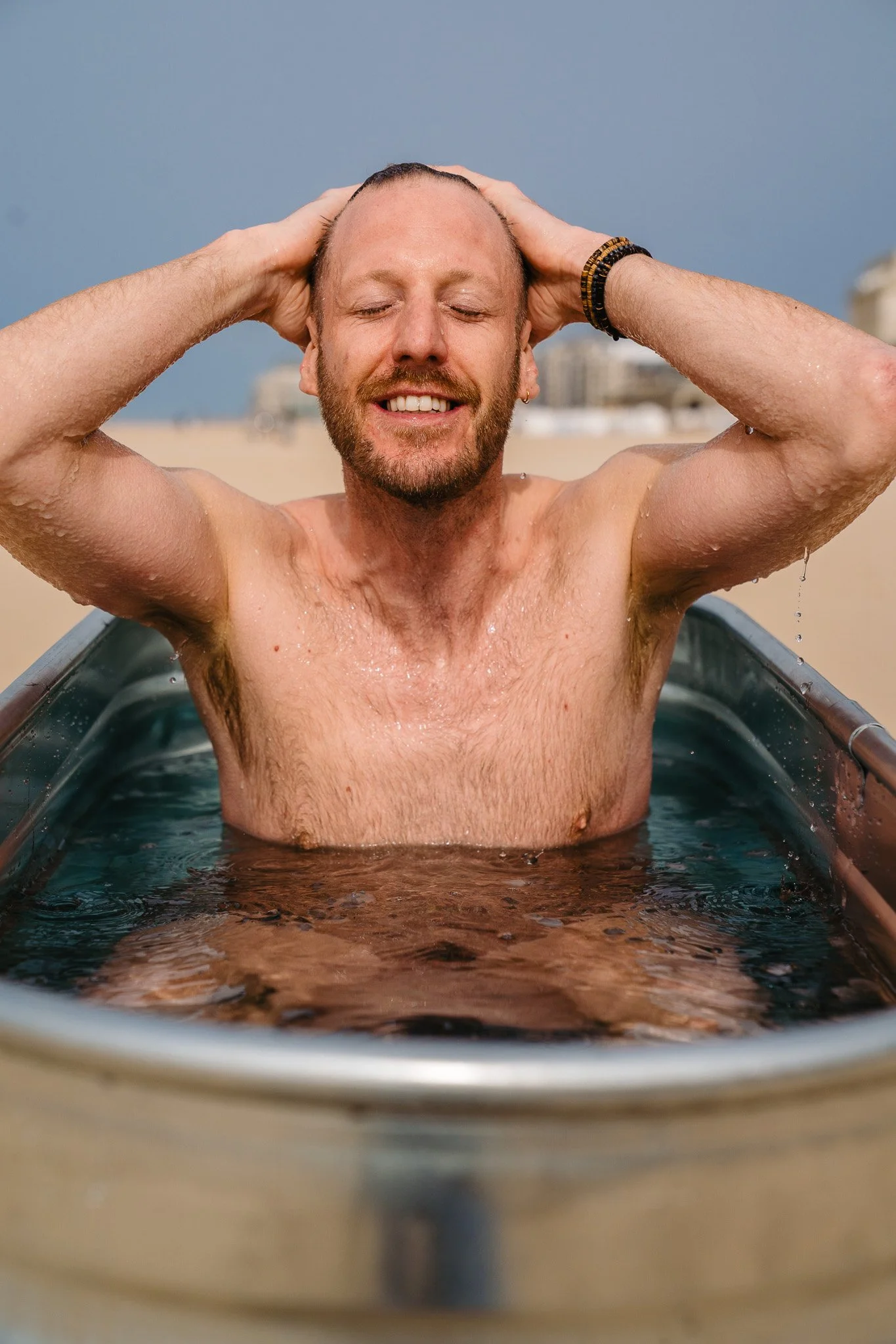 A man smiling with eyes closed, sitting in a Stock tank pool  filled with water on a beach, with his hands holding his head, and buildings in the background.
