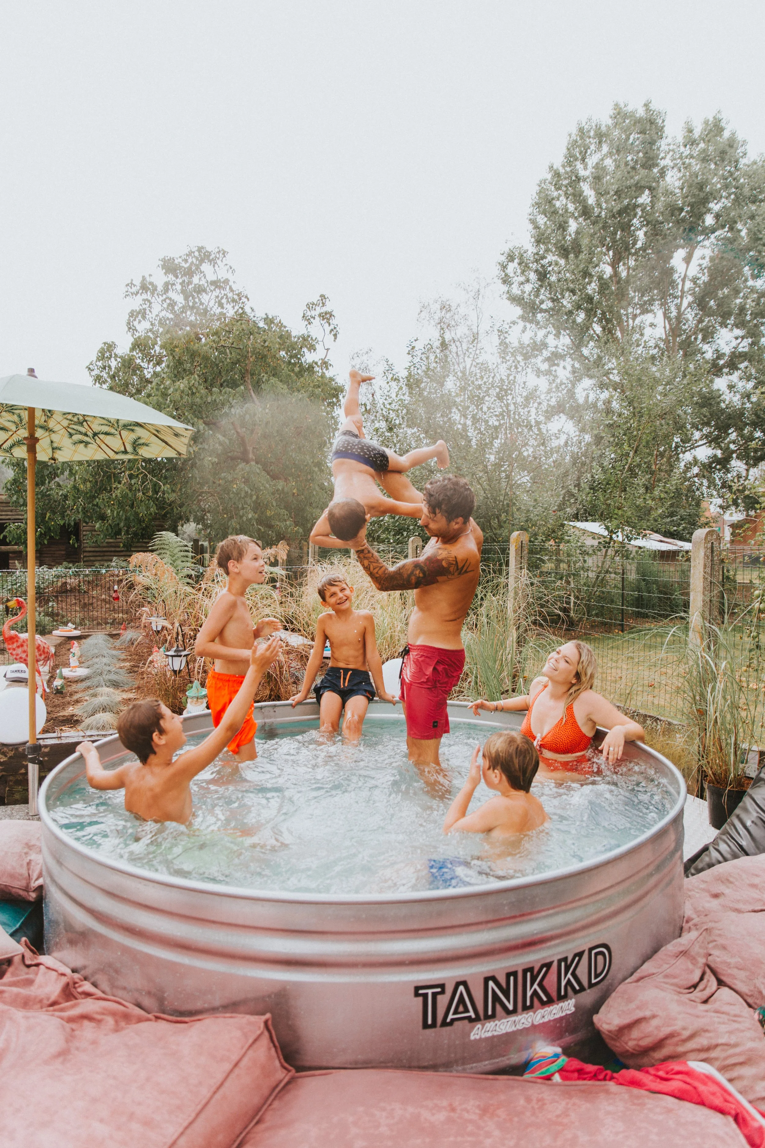 A group of kids and a man in a backyard hot tub playing and having fun, with one boy being lifted into the air by the man.