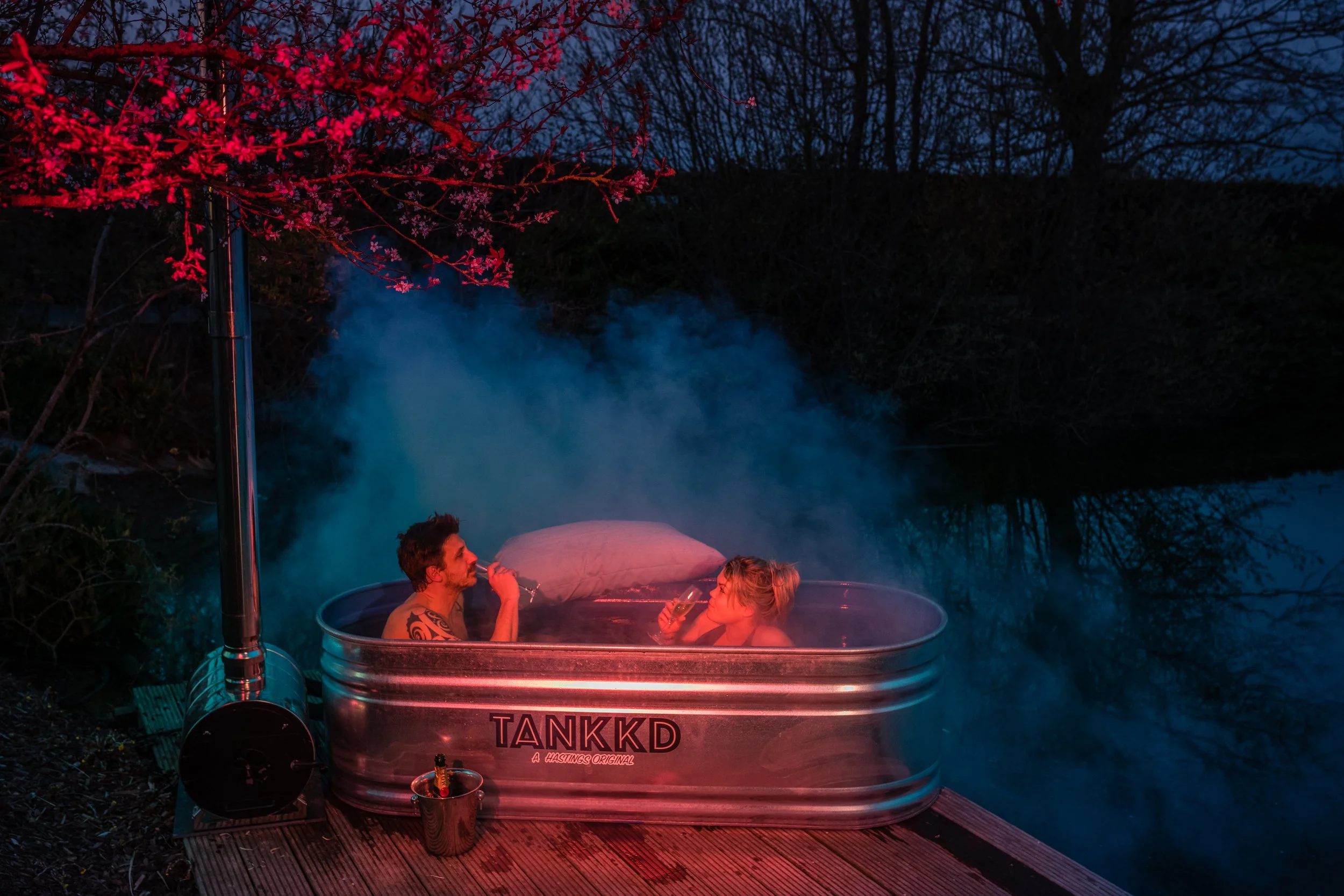 Two people relaxing in a hot tub outdoors at dusk, with steam rising in a natural setting, illuminated by pink and blue lighting.