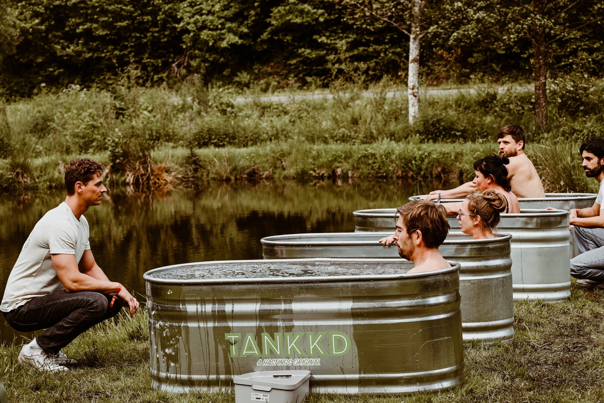 People sitting in Ice baths arranged in a line outdoors near a river, facing a man crouching in front of the first tub, amidst green trees and grass.