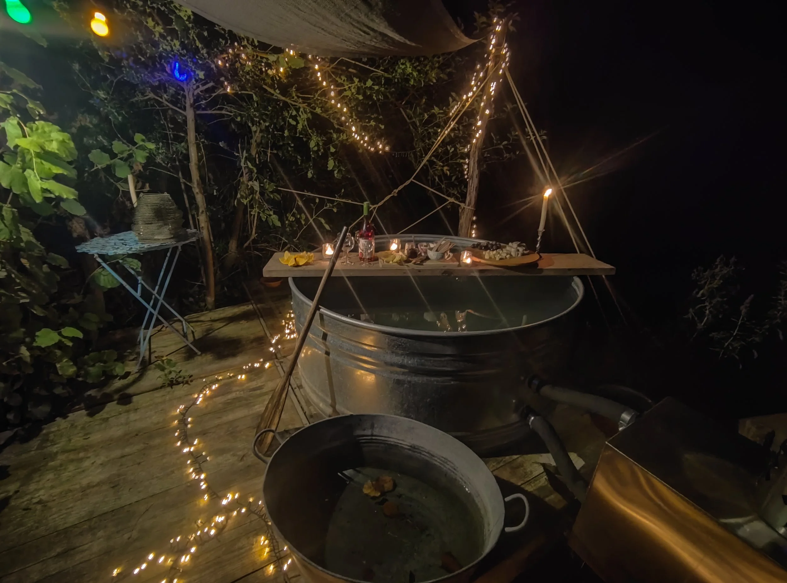 Outdoor nighttime scene with a hot tub surrounded by string lights and candles, a small table with a bottle and snacks, and a wooden deck with a metal bucket and a grill.