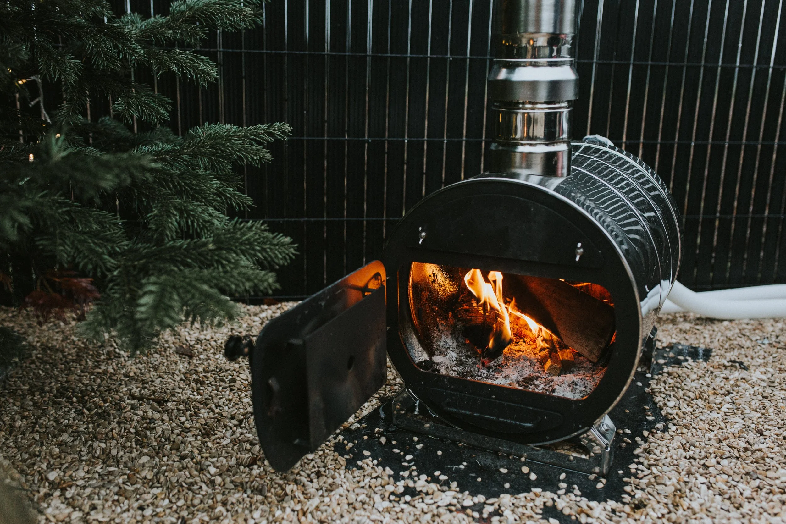 A small black wood stove with an open door, showing a fire inside, situated on gravel with a green pine branch on the left and a black fence in the background.