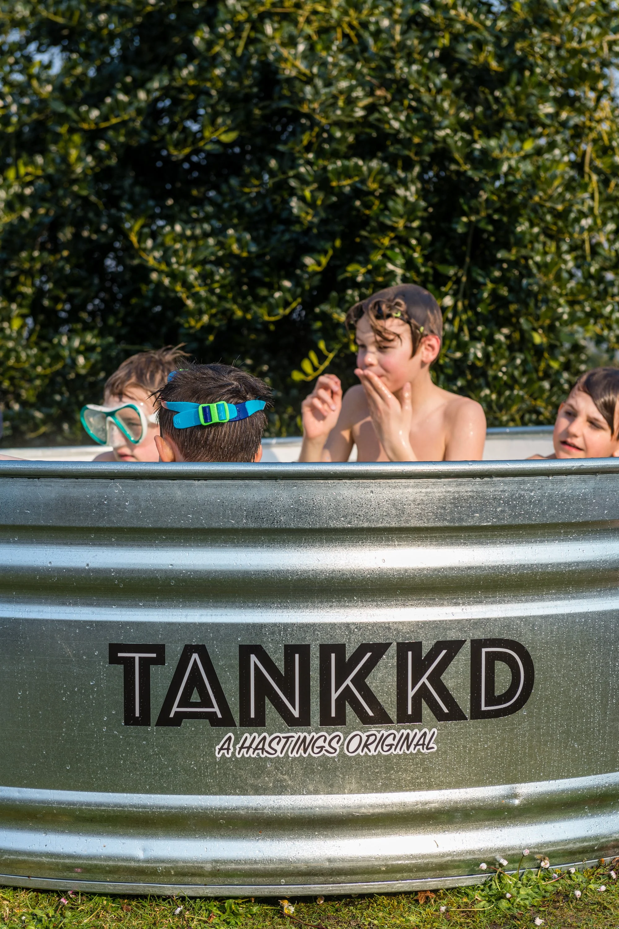 Four children playing in a metal tub labeled 'TANKKD: A Hastings Original' outdoors on a sunny day, with three boys visible and one wearing goggles.