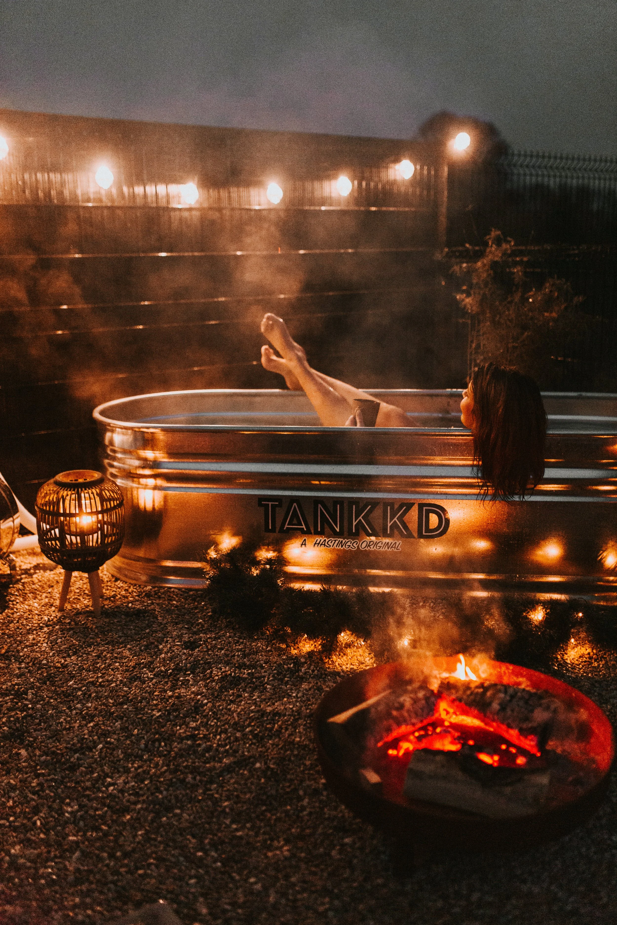 A person relaxing in a hot tub outdoors at night, with string lights above and a fire pit nearby.