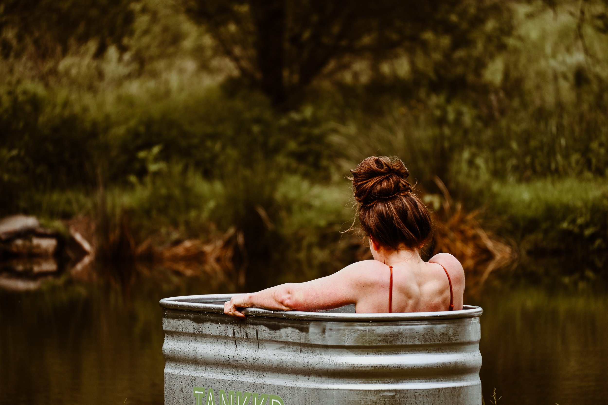 A woman with her hair in a bun sitting in a ice bath outdoors near a body of water, with lush green trees in the background.