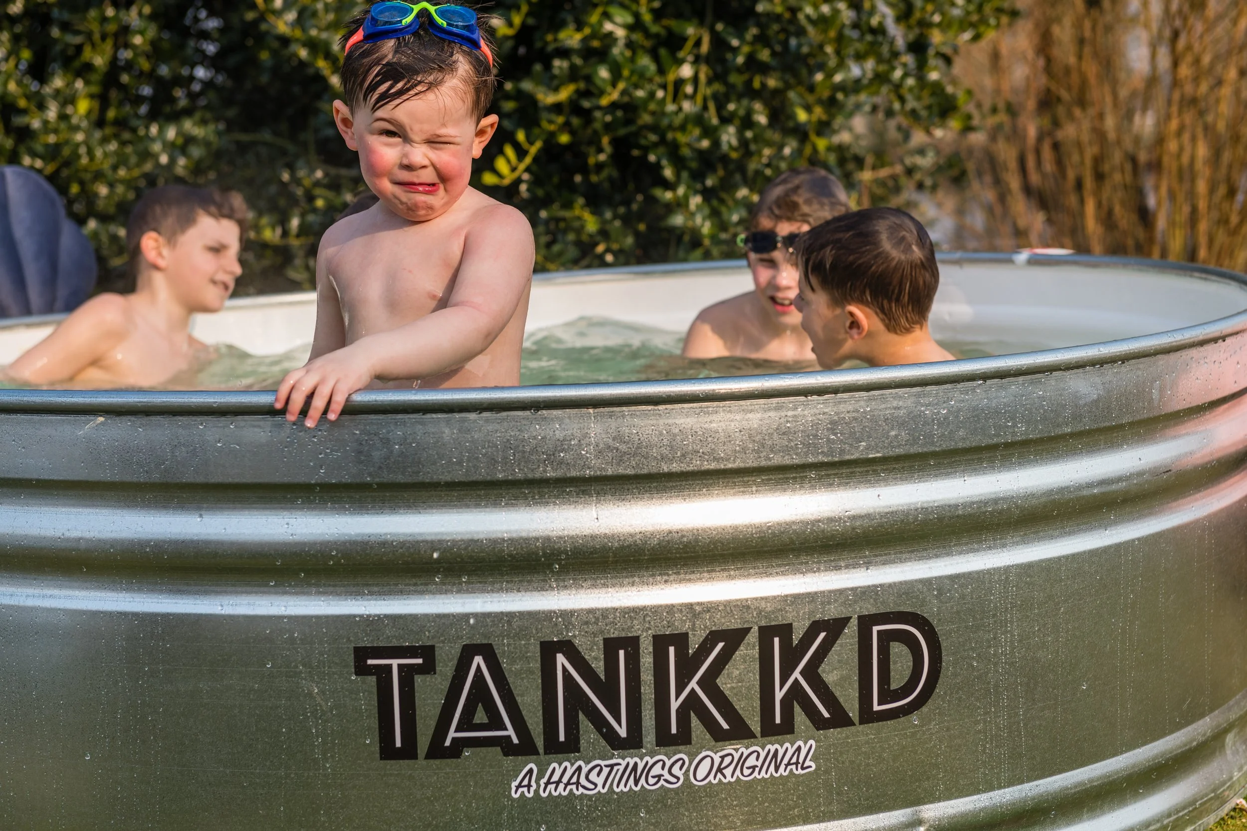 Four young boys playing and splashing in a metal kiddie pool outdoors on a sunny day, surrounded by trees and bushes.