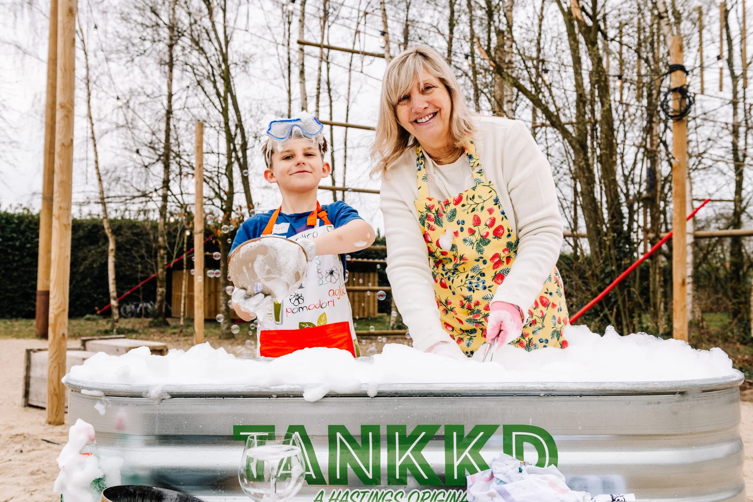 A smiling woman and a young boy stand behind a large filled with soap bubbles outdoors. The boy, wearing a blue shirt, an apron, and goggles, is holding a sponge. The woman is wearing a floral apron and pink gloves, leaning over the tub. There are so