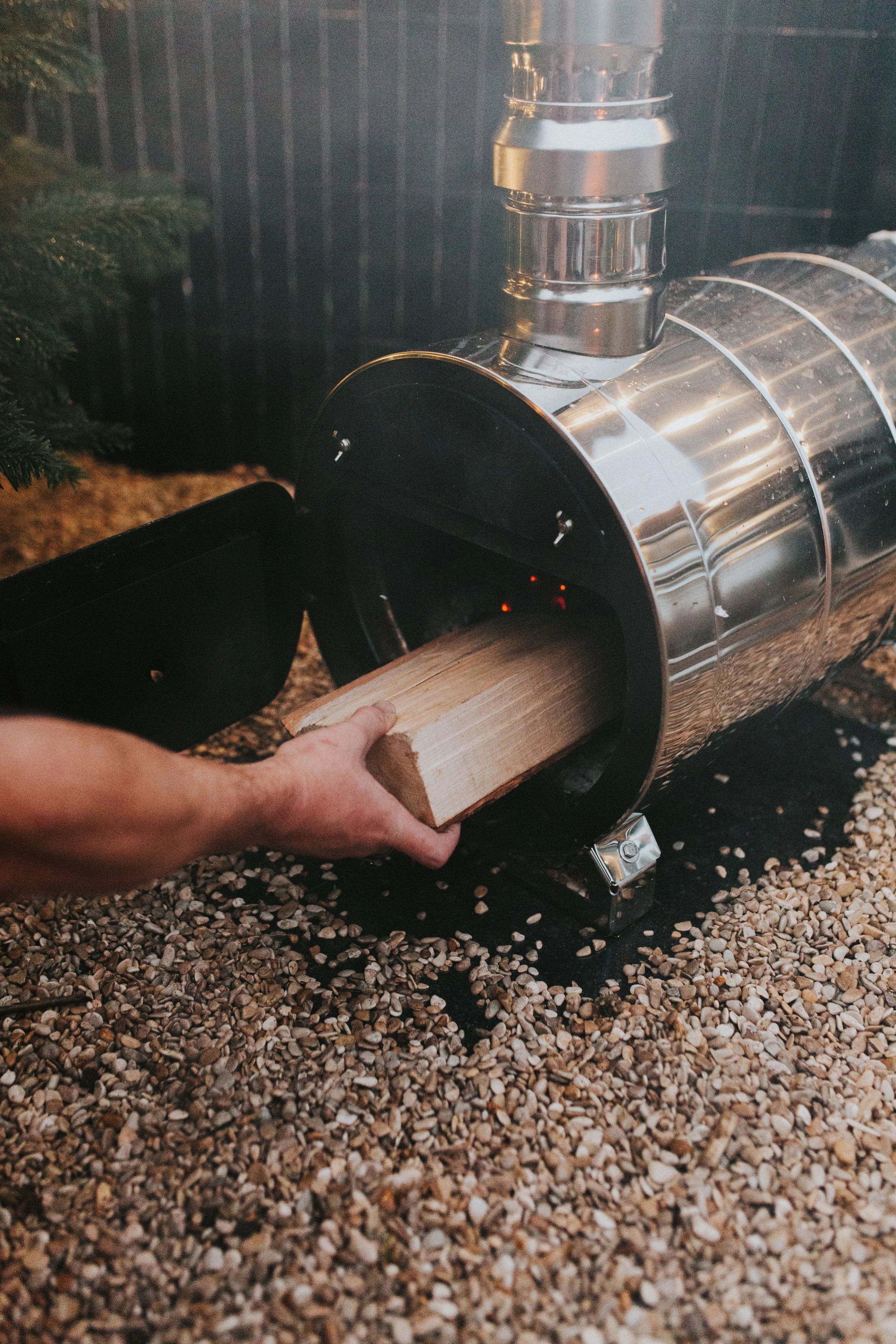 A person inserting a piece of firewood into a metal wood furnace with a vent pipe, surrounded by pebbles.