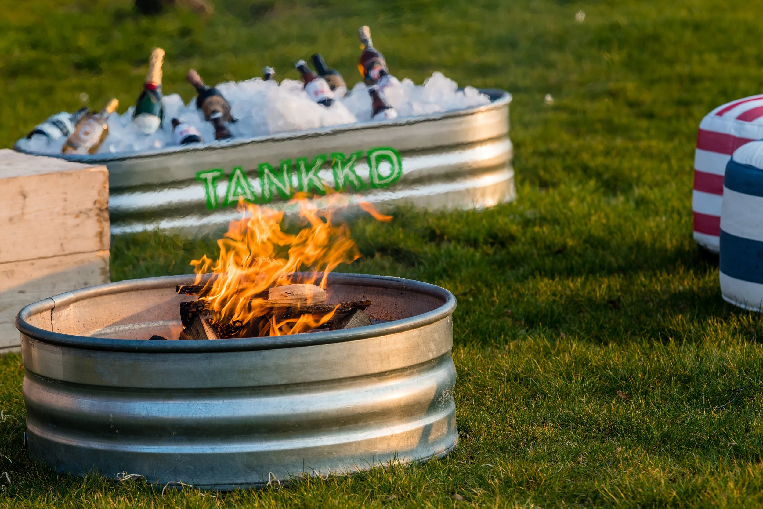 A fire pit with burning wood in the foreground, and a metal tub filled with ice and bottles of drinks in the background.