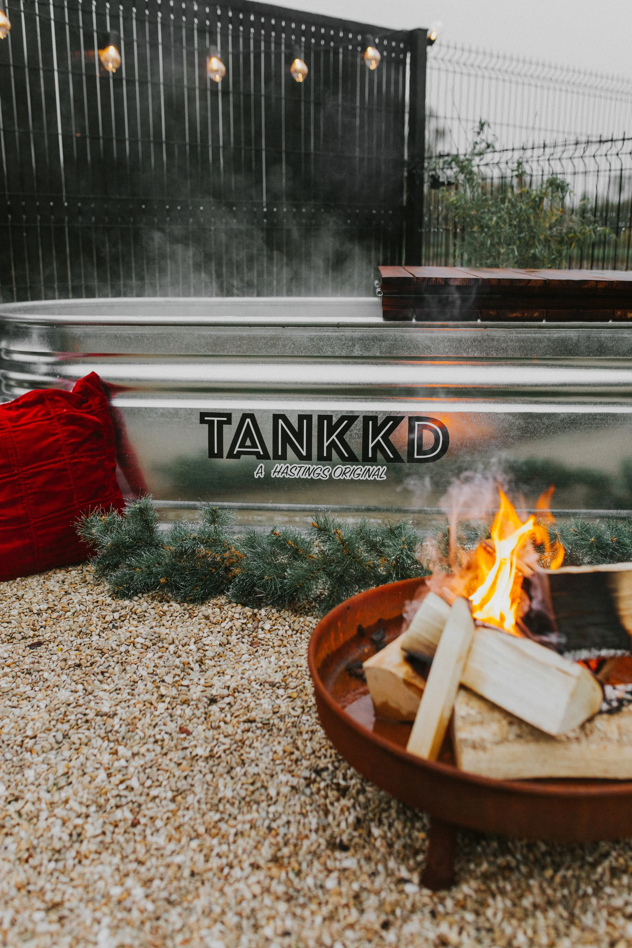 A metal hot tub with string lights above it, labeled 'TANKKD A HASTINGS ORIGINAL'. In front of the hot tub, there is a small fire pit with burning wood, and a red pillow is resting on the gravel ground next to a sprig of pine. A black fence surrounds